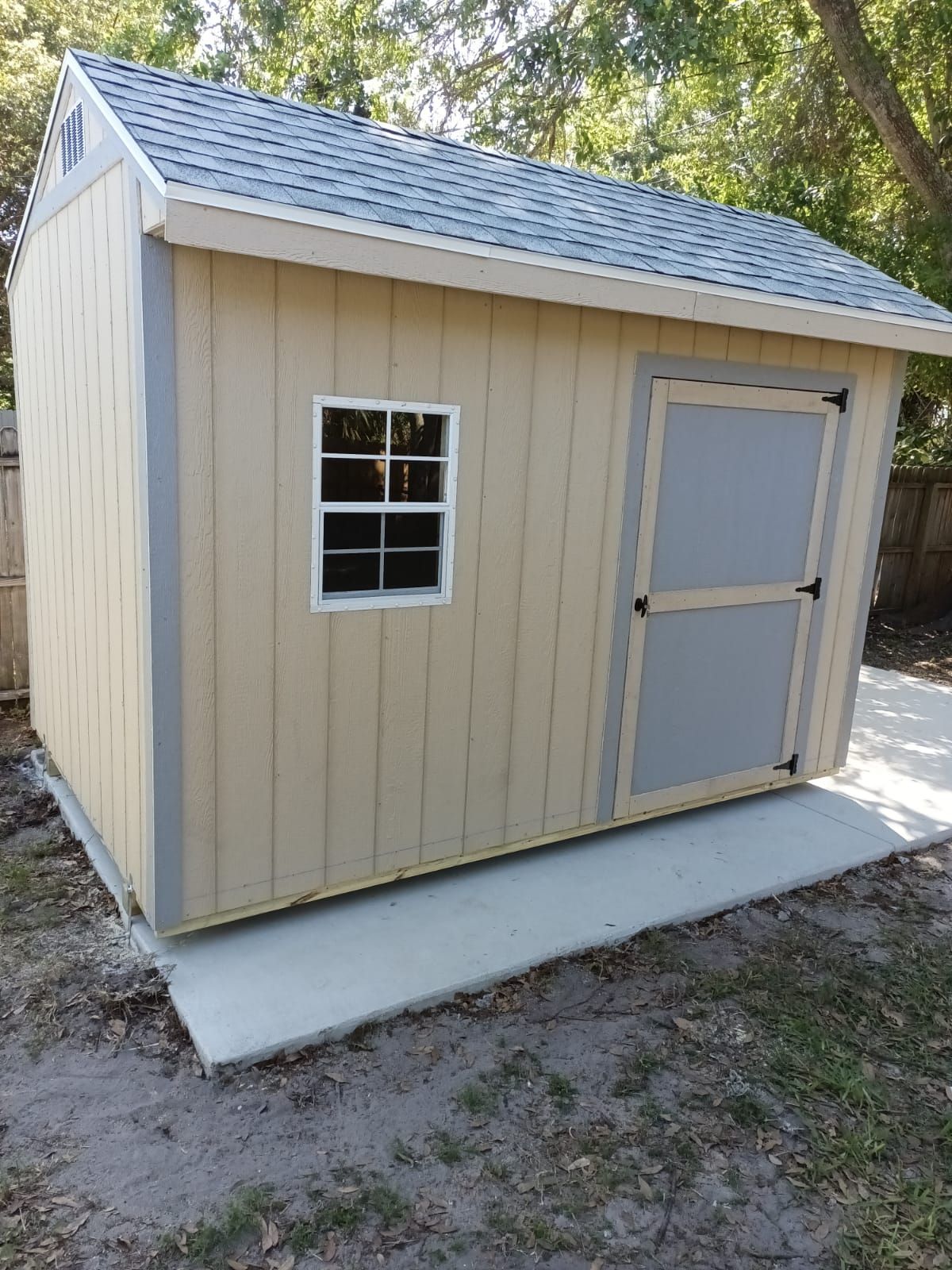 A small shed with a window and a gray door is sitting on the side of a driveway.