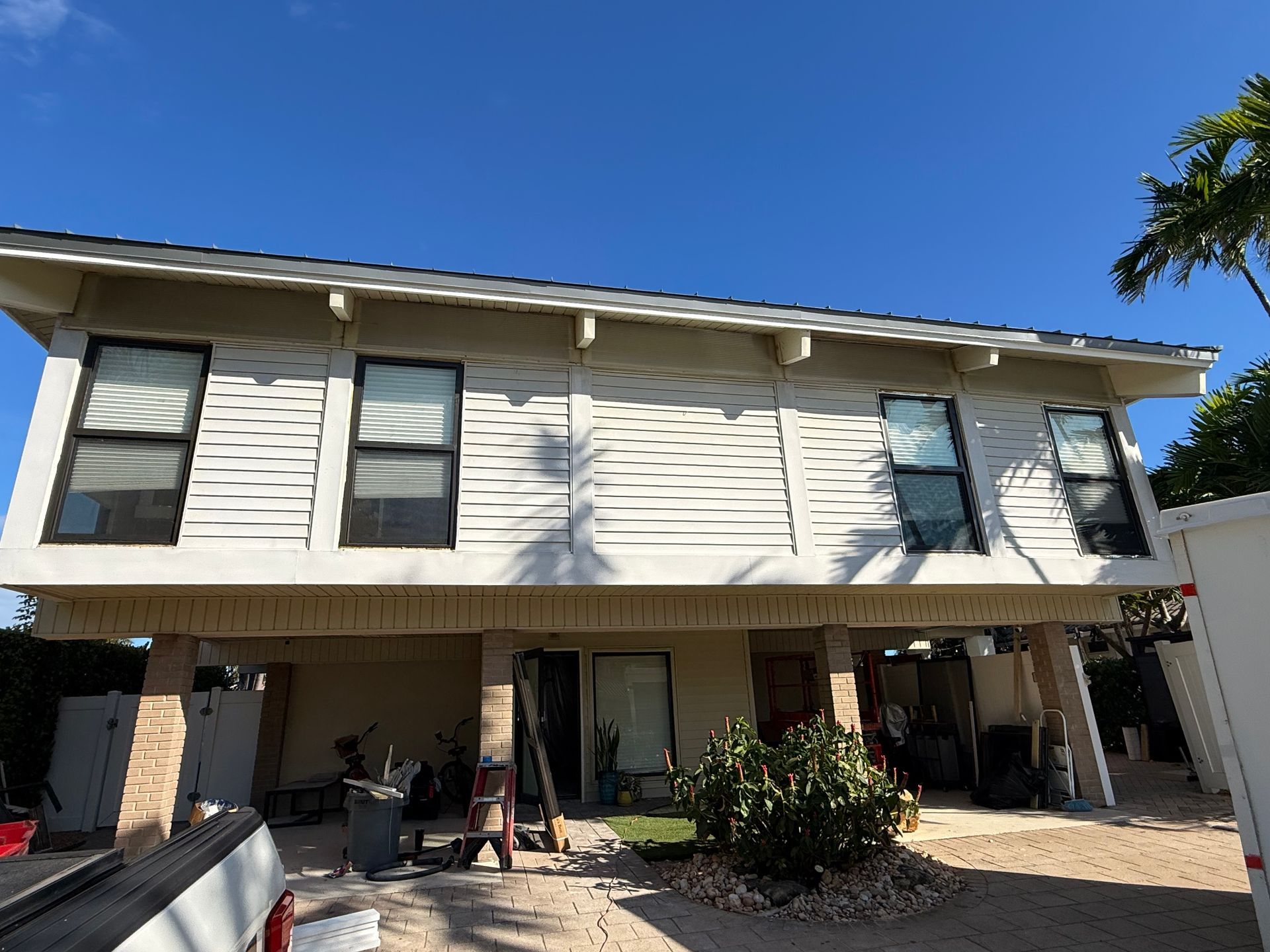 Two-story light-colored house elevated on stilts with multiple windows under a bright blue sky.