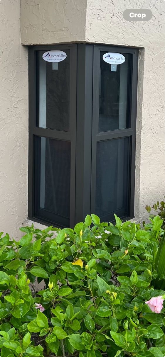 A dark gray corner window with a screen is set in a beige wall and surrounded by green shrubbery.