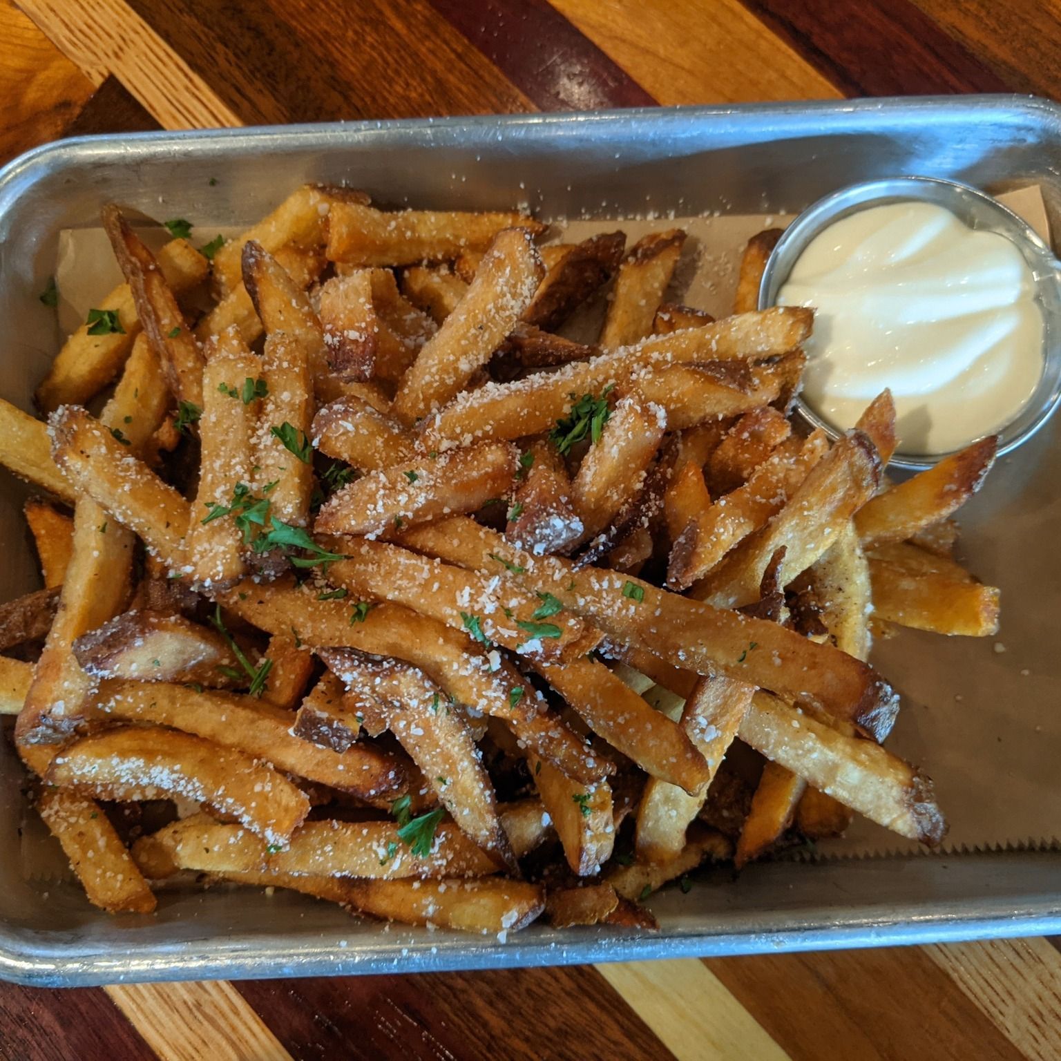 Tray of golden fries with grated cheese and a small cup of dipping sauce on a wooden table