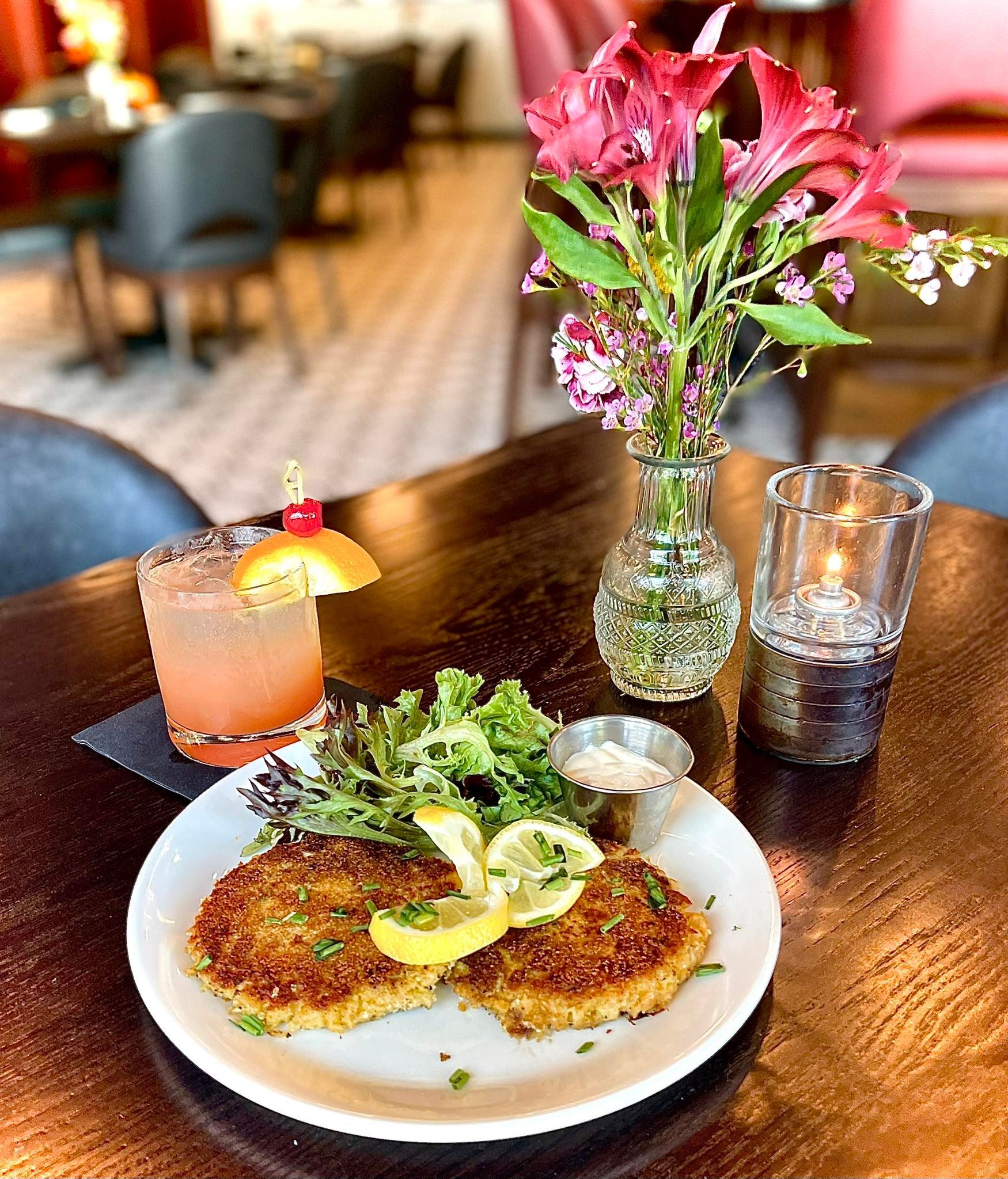Brunch plate with pancakes, greens, and a cocktail on a wooden table beside flowers and a candle holder