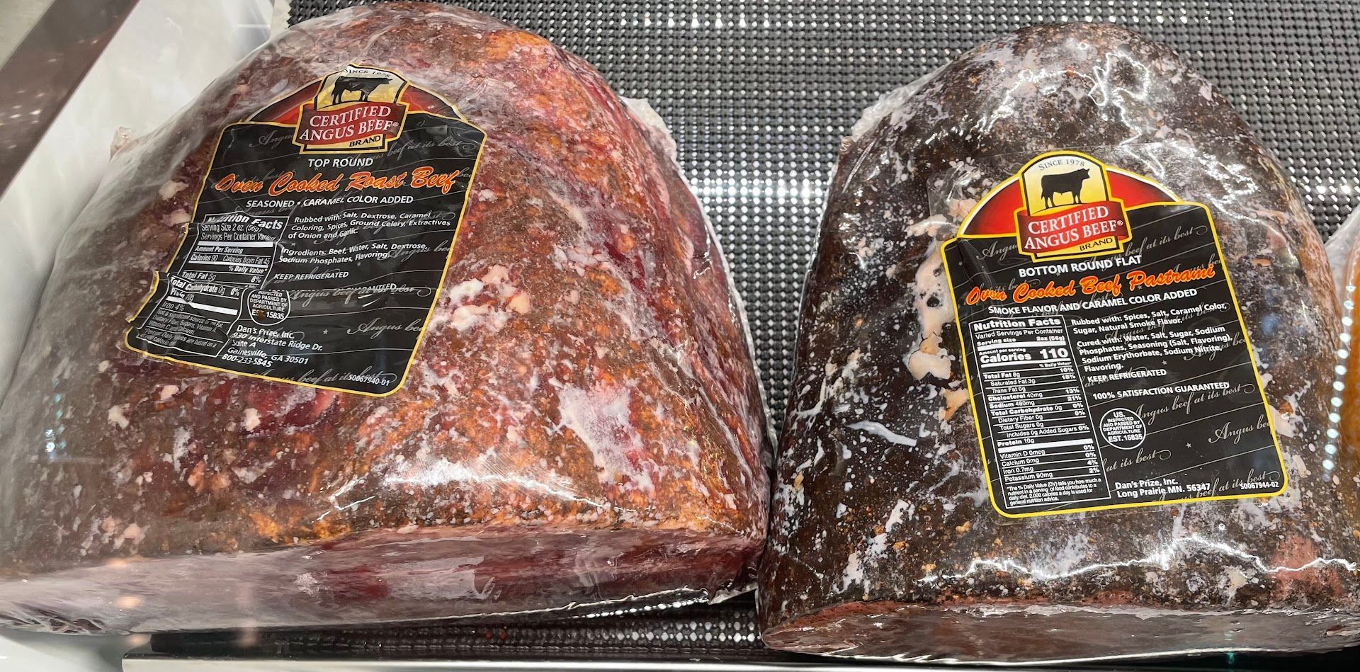 Two wrapped cured meats on a refrigerated display shelf, with black labels and white frost.