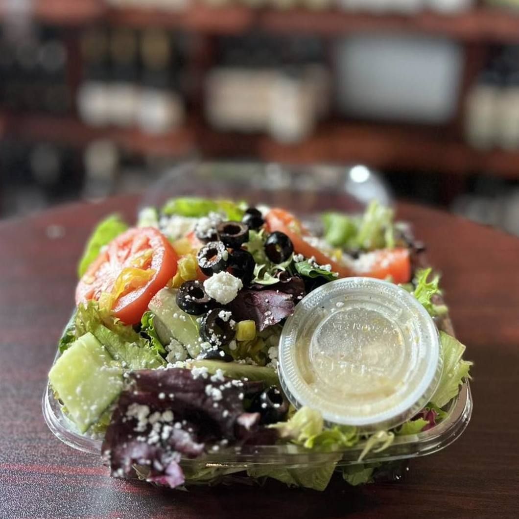 Clear plastic container of Greek salad with lettuce, tomato, olives, feta, and dressing cup on a table
