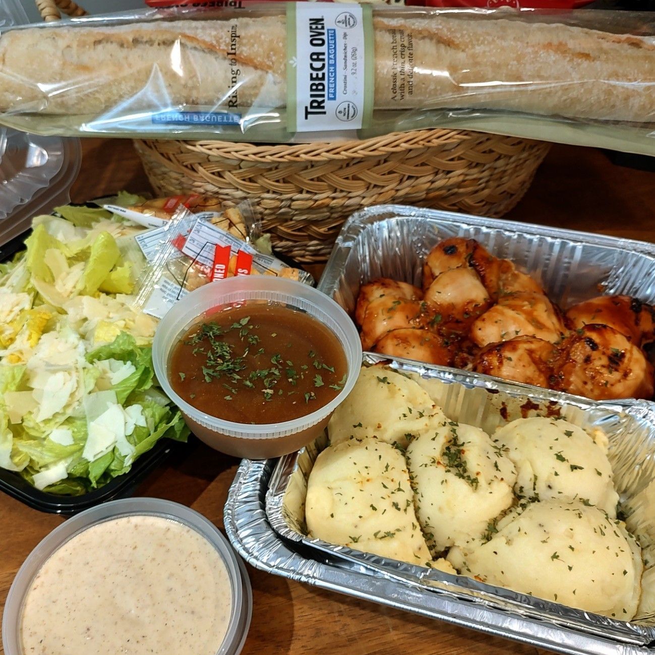 Takeout meal with salad, chicken, potatoes, two sauces, and a baguette in the background