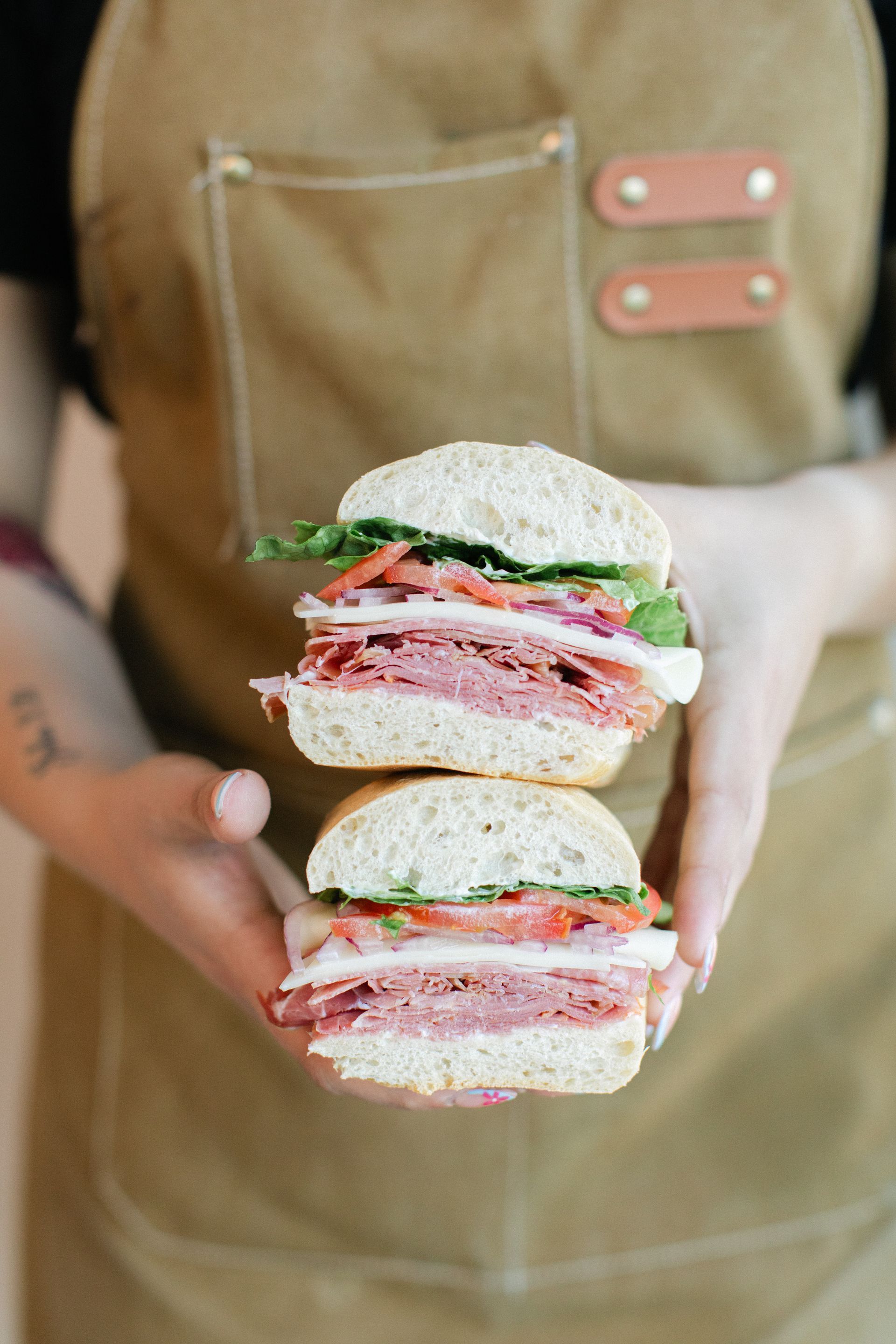 Person holding a stacked sandwich with meat, lettuce, and tomato in a kitchen apron
