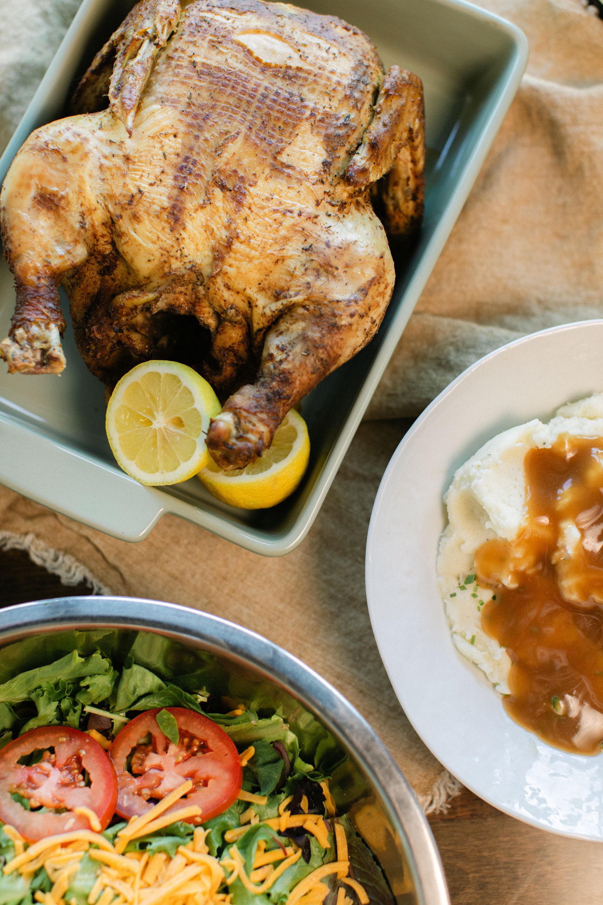 Roasted whole chicken with lemon wedges, salad, and a bowl of gravy on a table