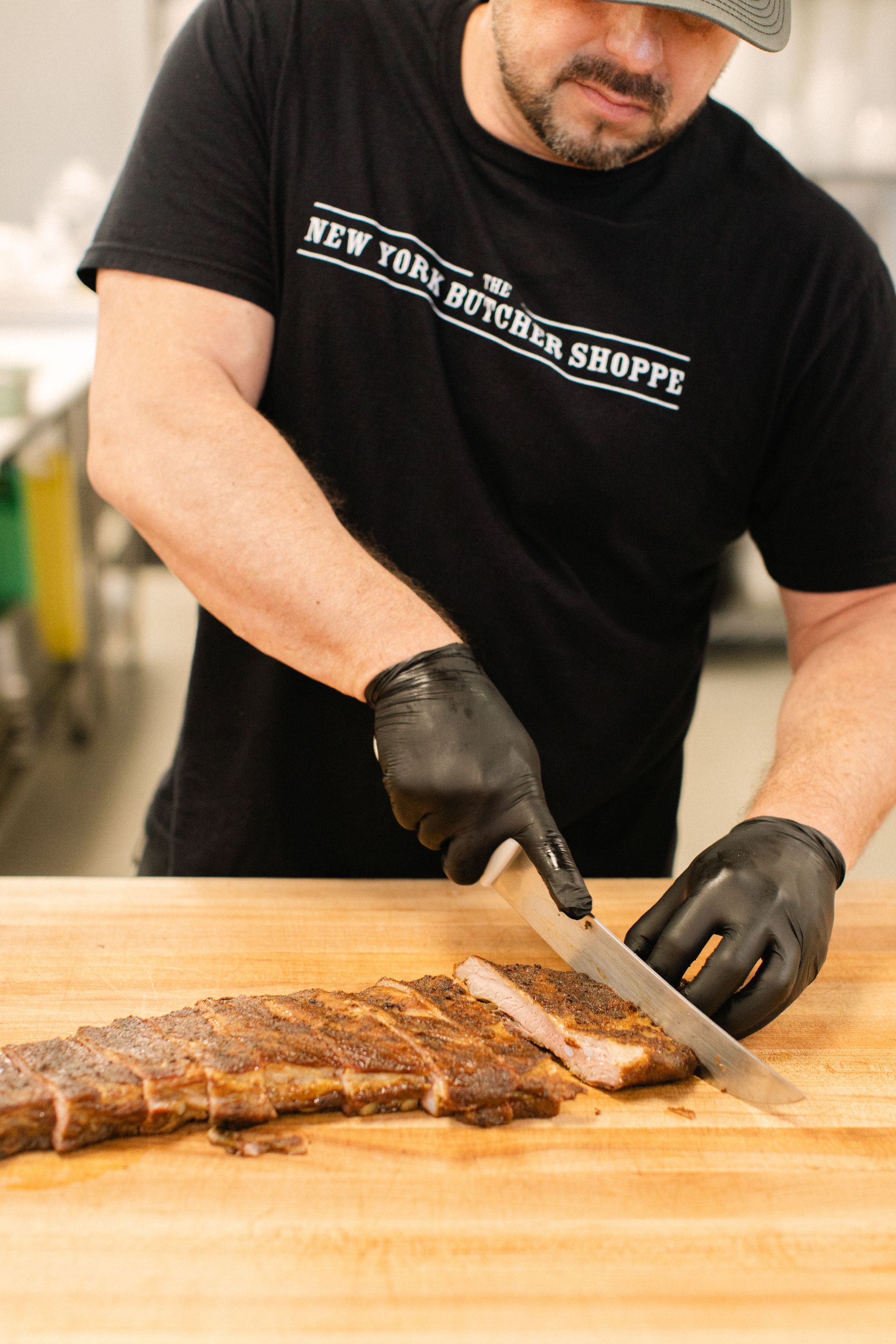 Man slicing cooked meat on a wooden cutting board with a knife, wearing black gloves and a black T-shirt