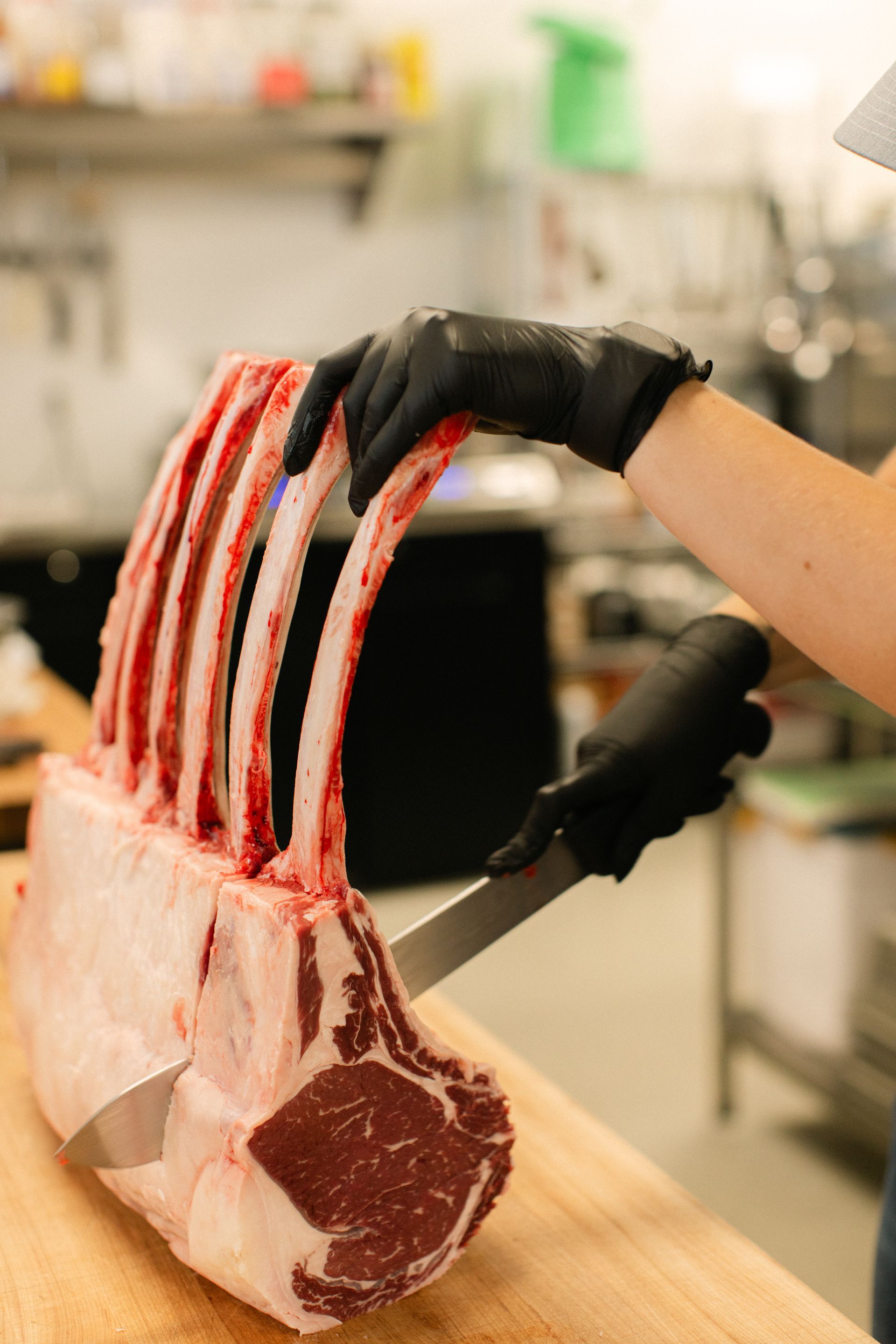A gloved hand holds a rack of raw lamb ribs on a butcher’s cutting board.