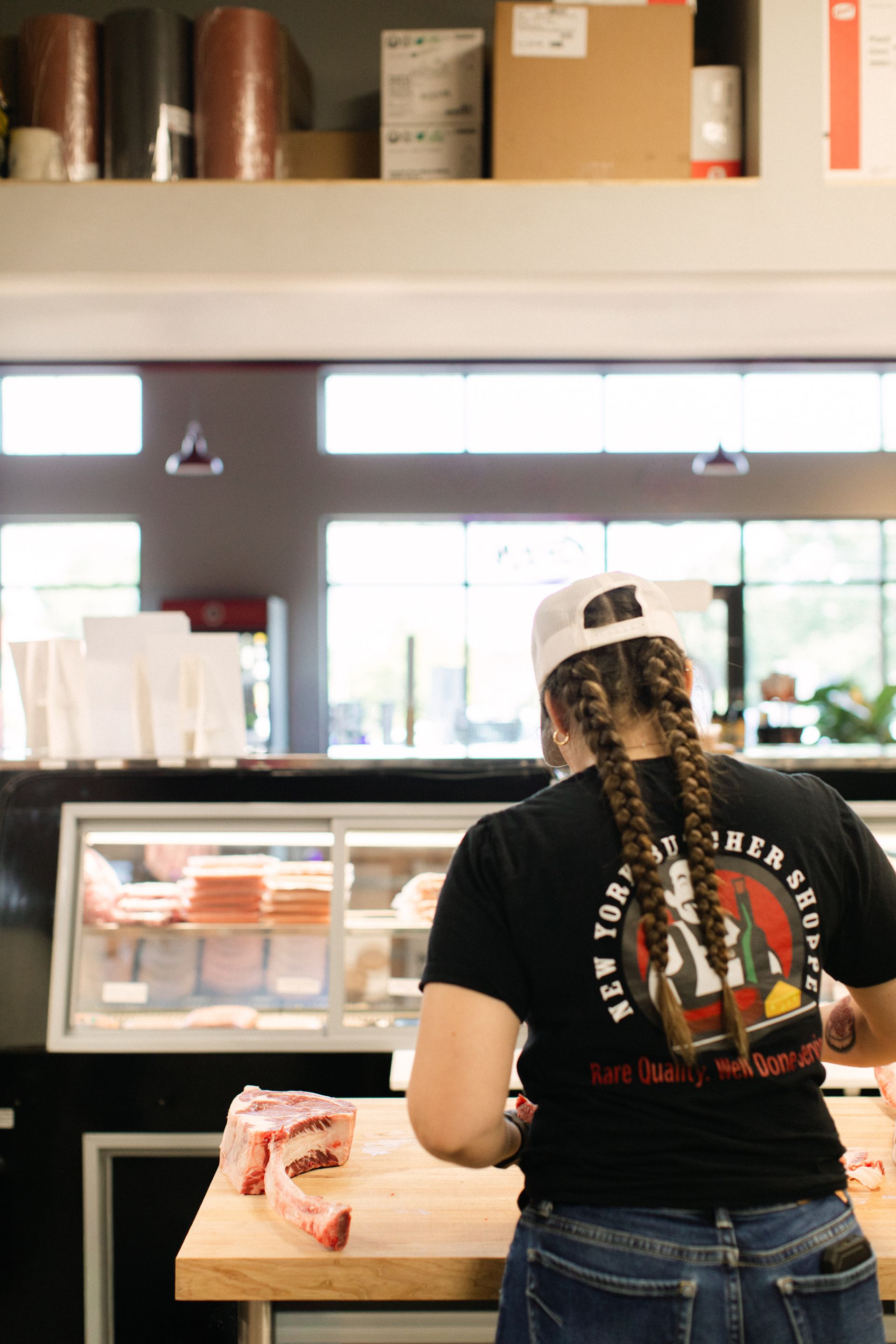 Person standing at a deli counter with braids, wearing a black T-shirt and white cap in a bright shop