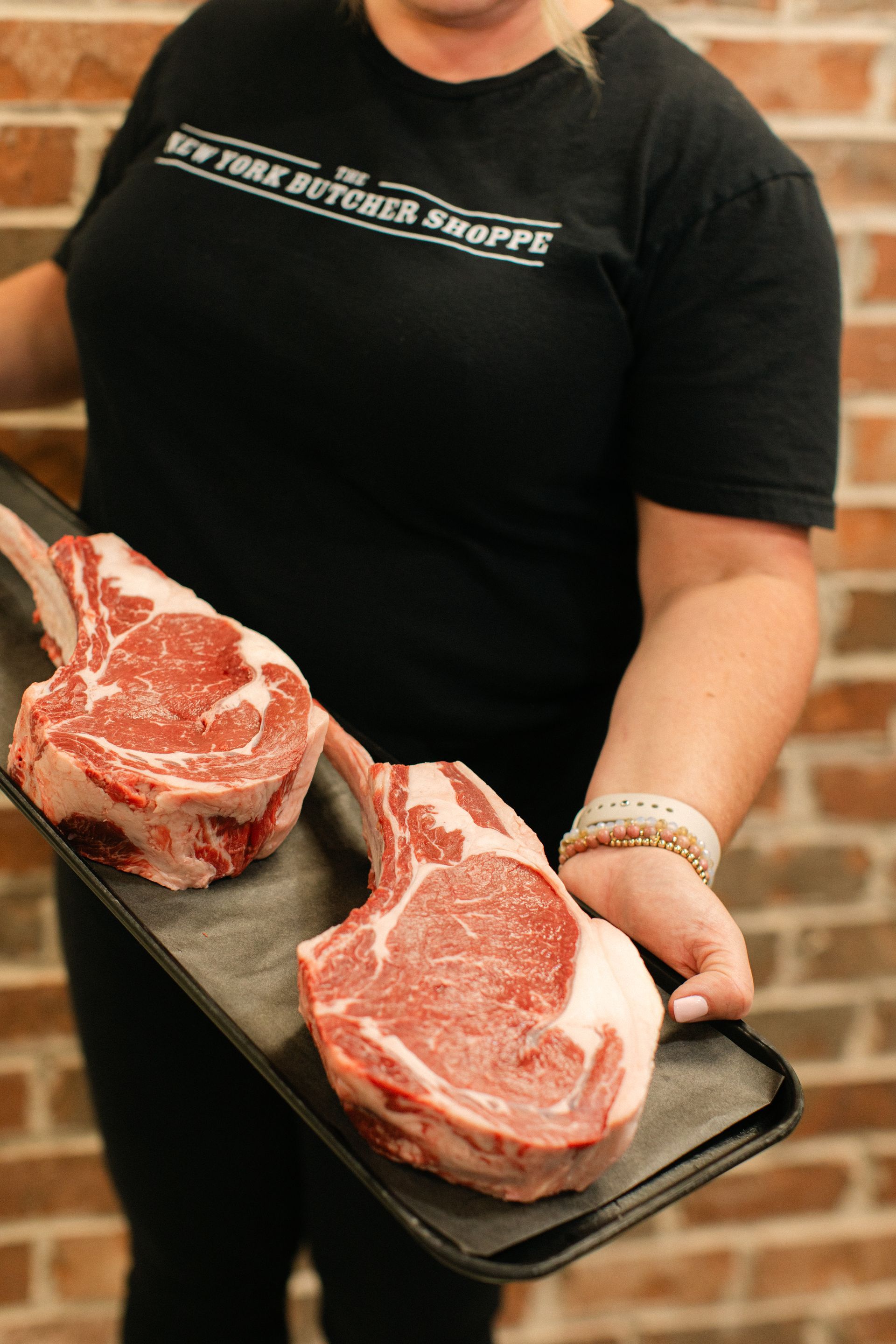 Person holding two raw ribeye steaks on a tray in front of a brick wall