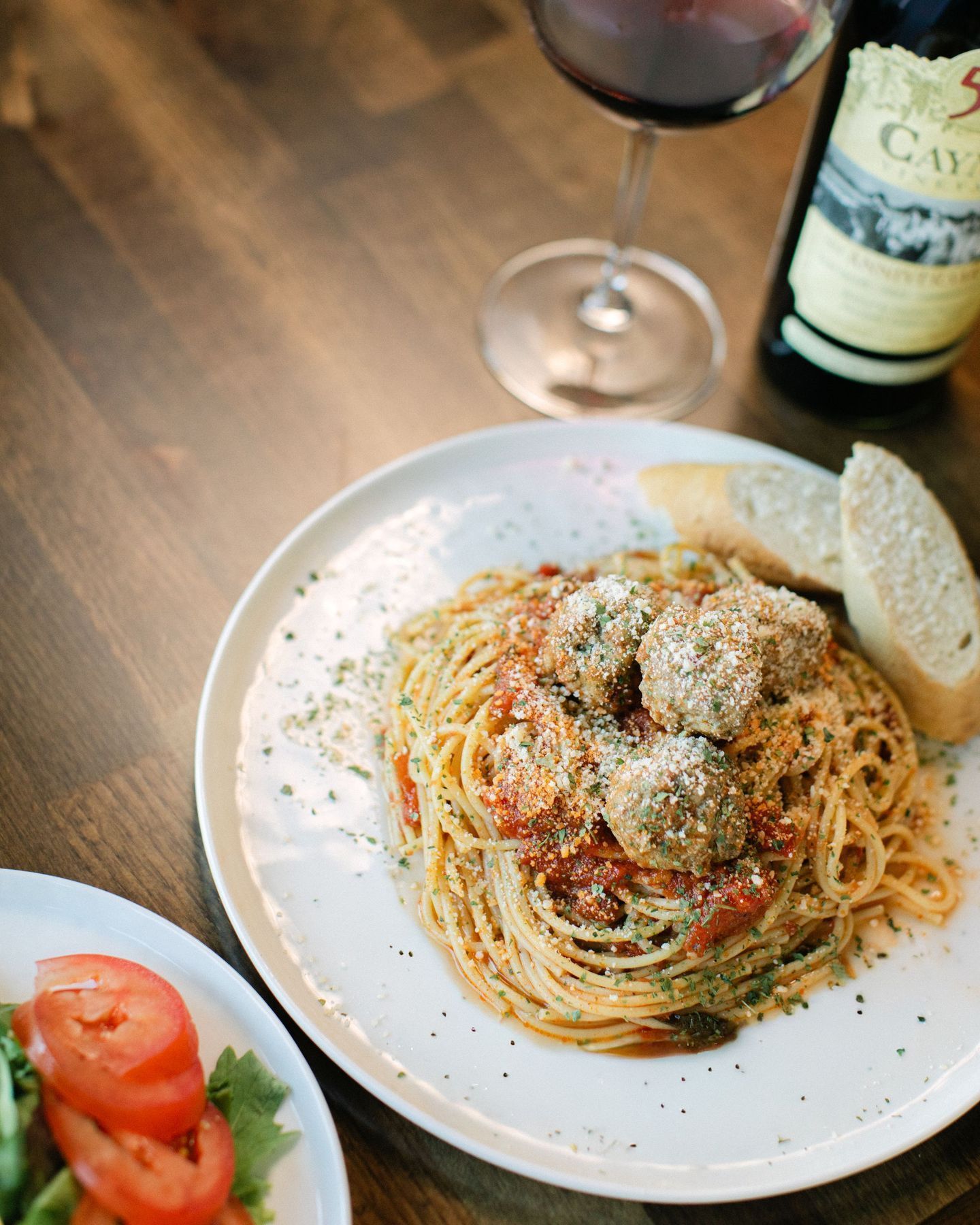 Plate of spaghetti with grated cheese and bread on a table, with a wine glass and bottle in the background