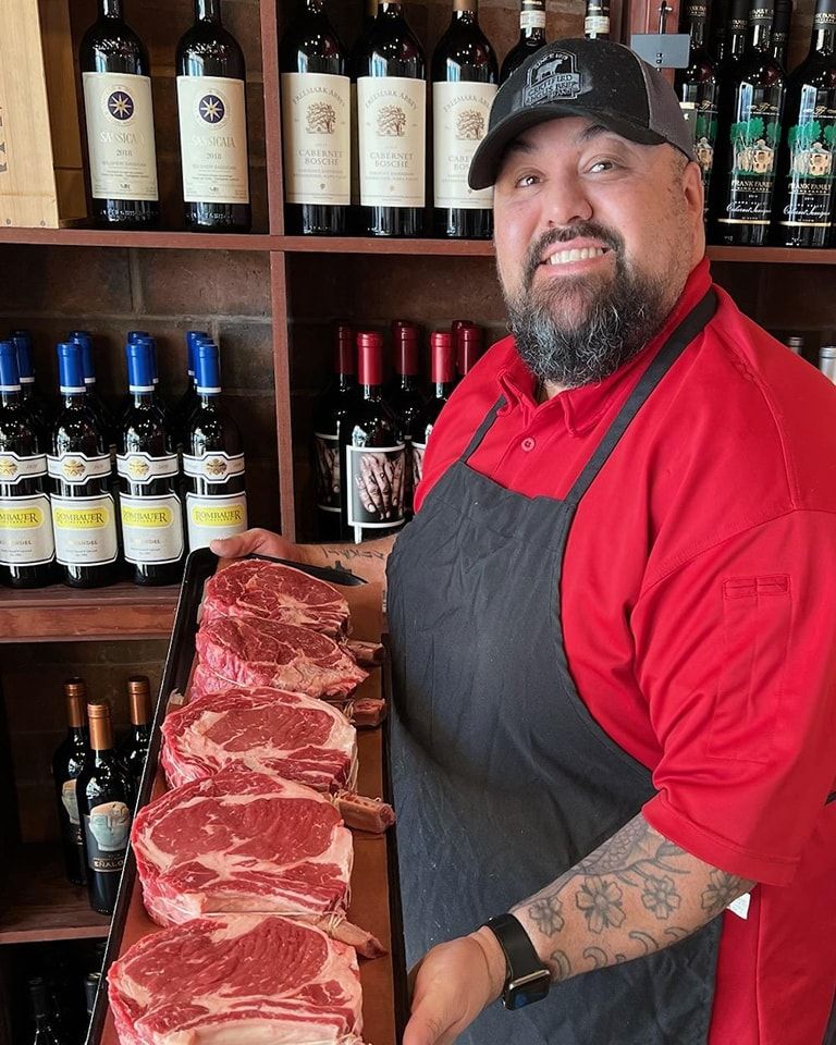 Butcher in red shirt and apron holding tray of raw steaks in front of wine bottles on shelves