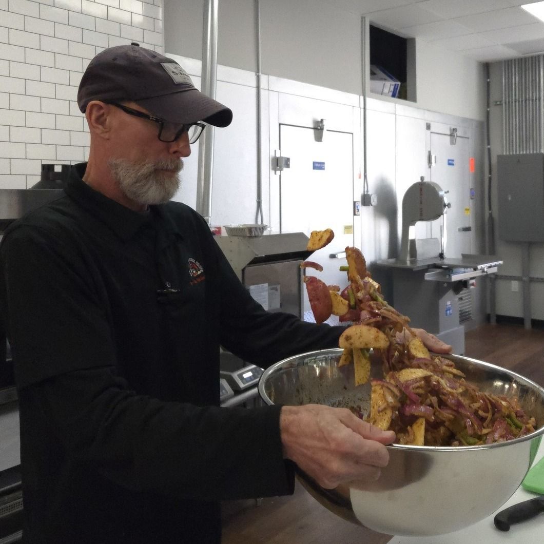 Man in black cap holding a large metal bowl of raw chicken in a kitchen