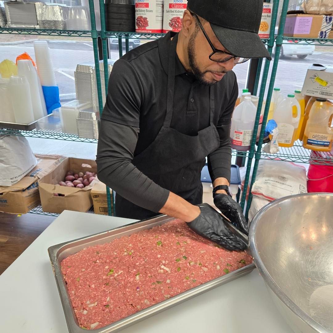 Person in a black apron and gloves preparing ground meat in a deli counter.