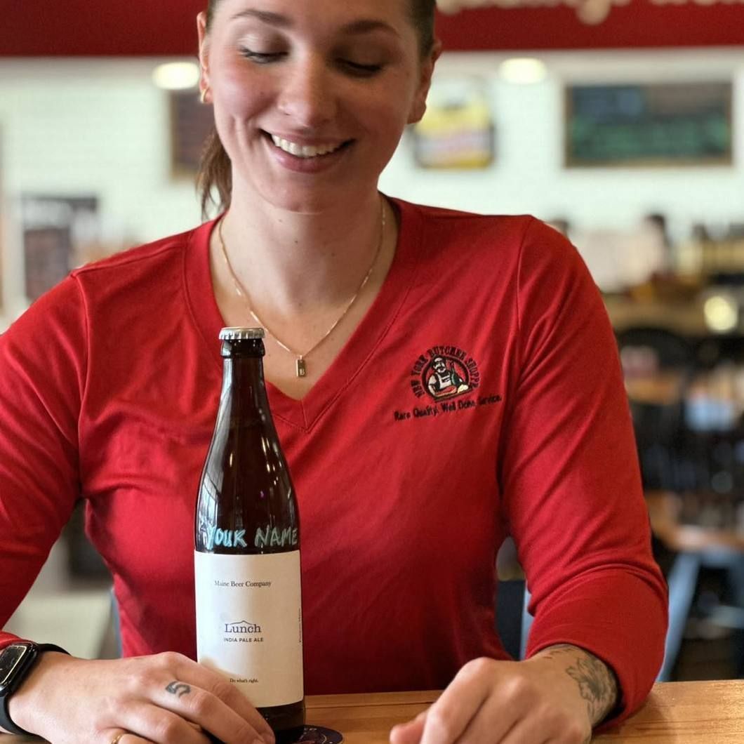 Smiling person in a red shirt holds a wine bottle at a table in a bright restaurant.