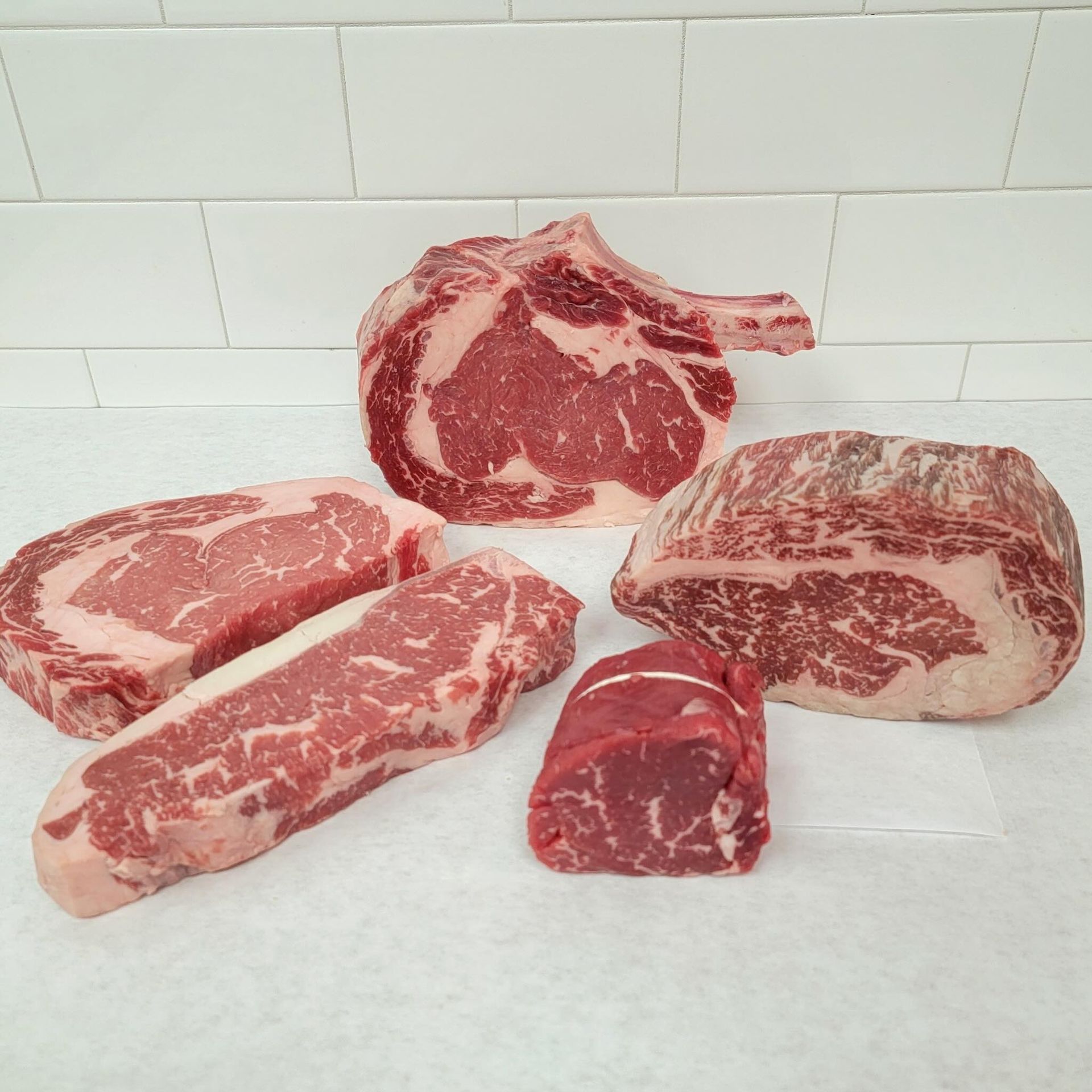 Raw marbled steaks and a rib roast arranged on a white countertop against a white tile backsplash
