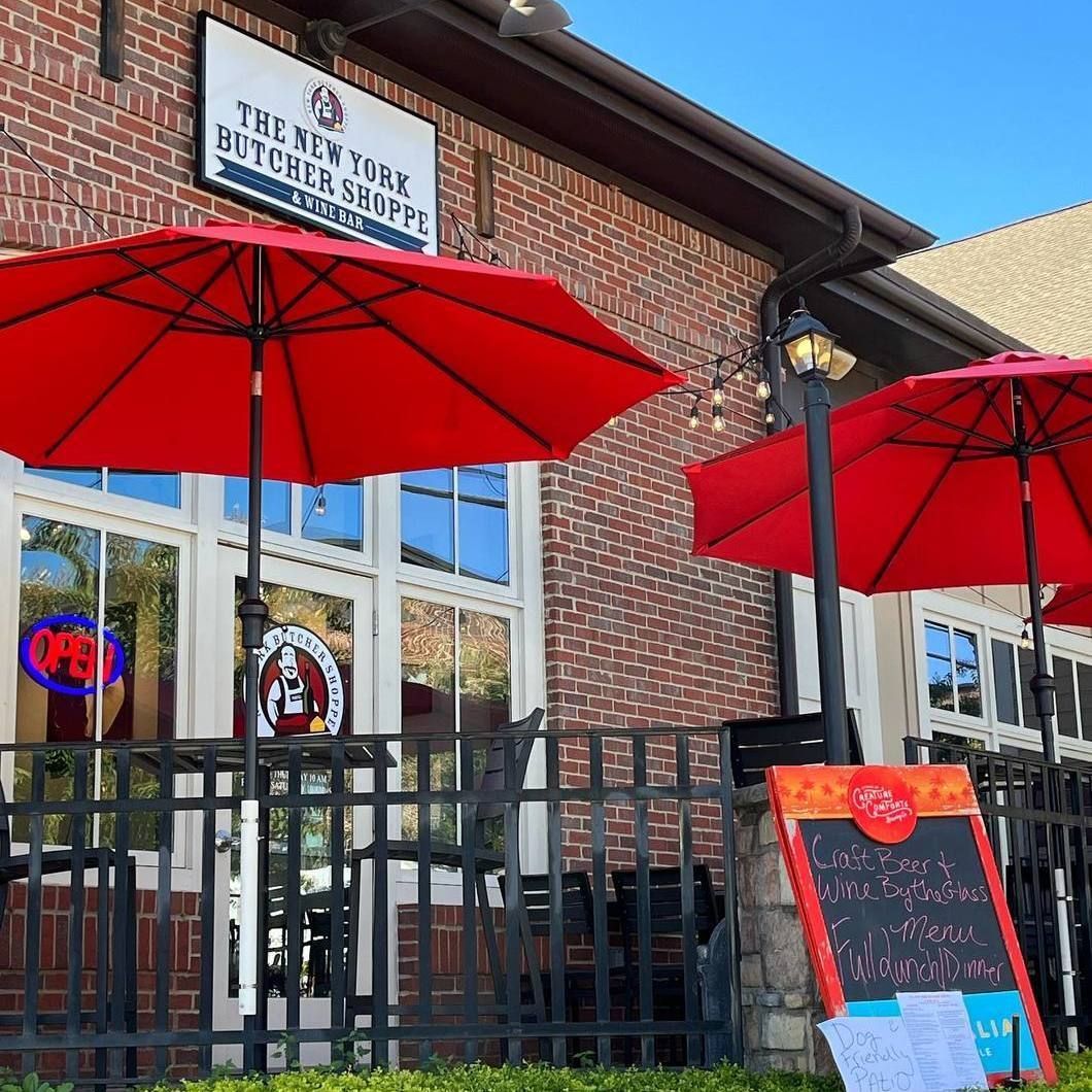 Brick storefront café with red umbrellas, black railing, and a sidewalk sign outside.