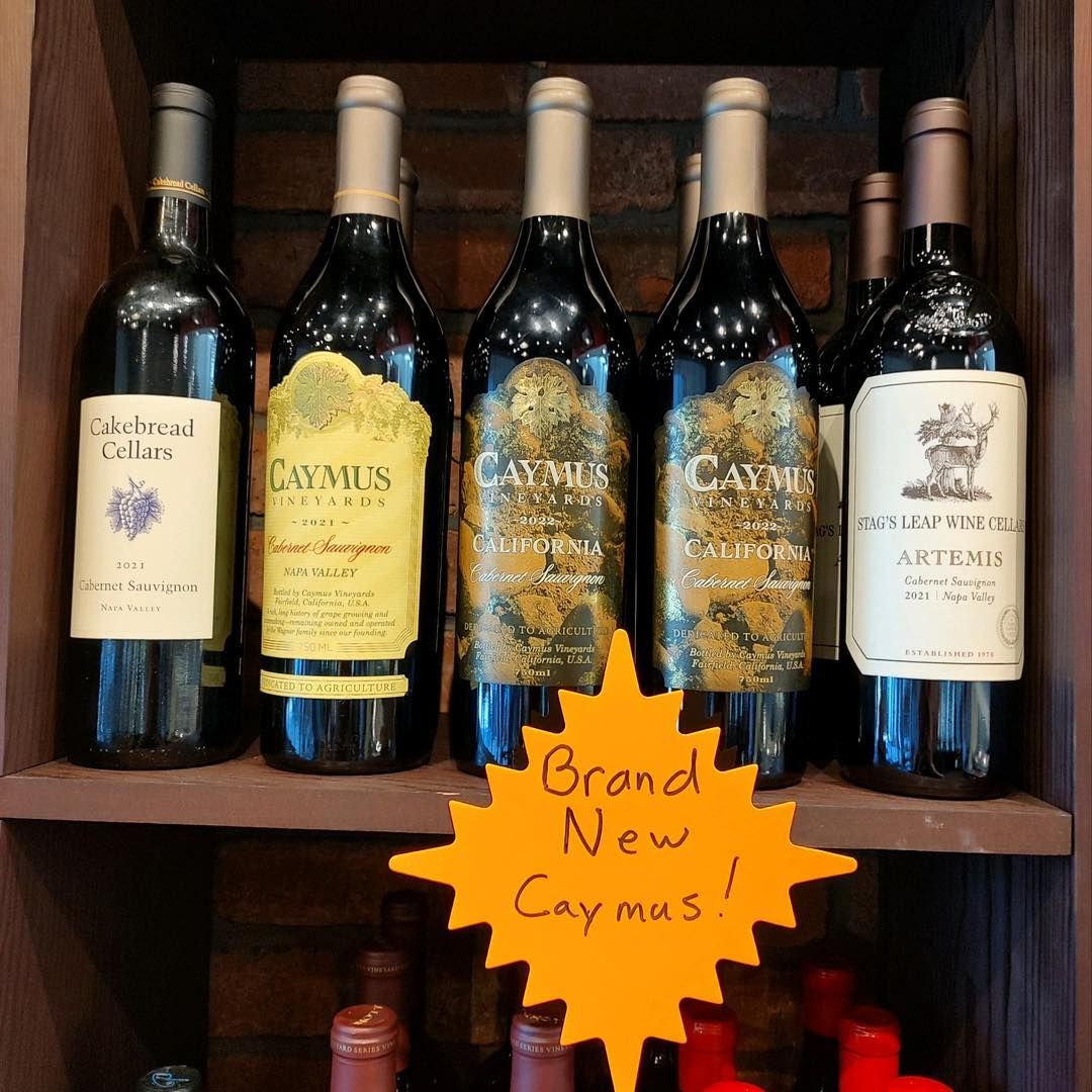 Five wine bottles on a shelf with a handwritten “Brand New Caymus” sign.