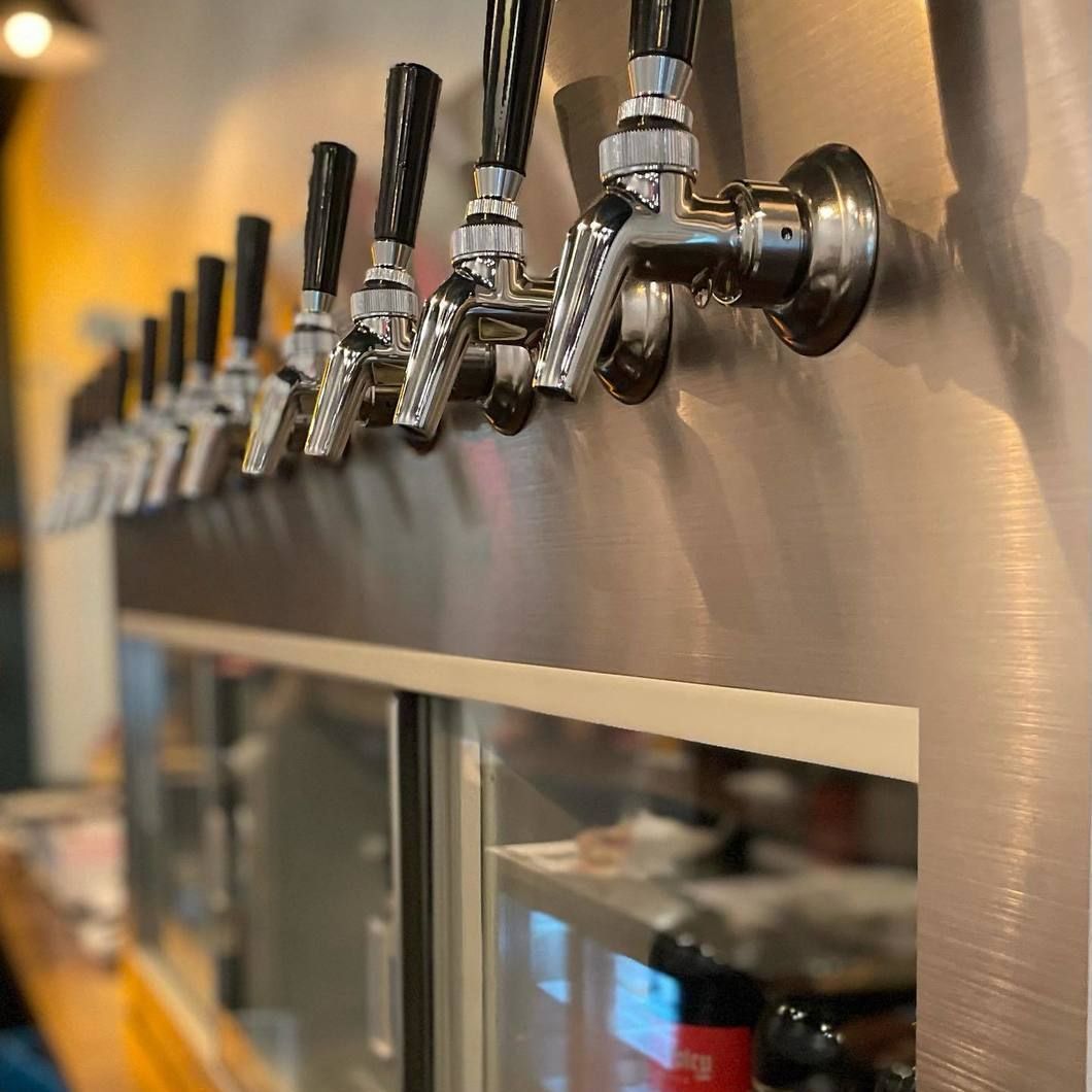 Row of chrome beer taps on a stainless-steel bar counter in a warm-lit brewery or pub