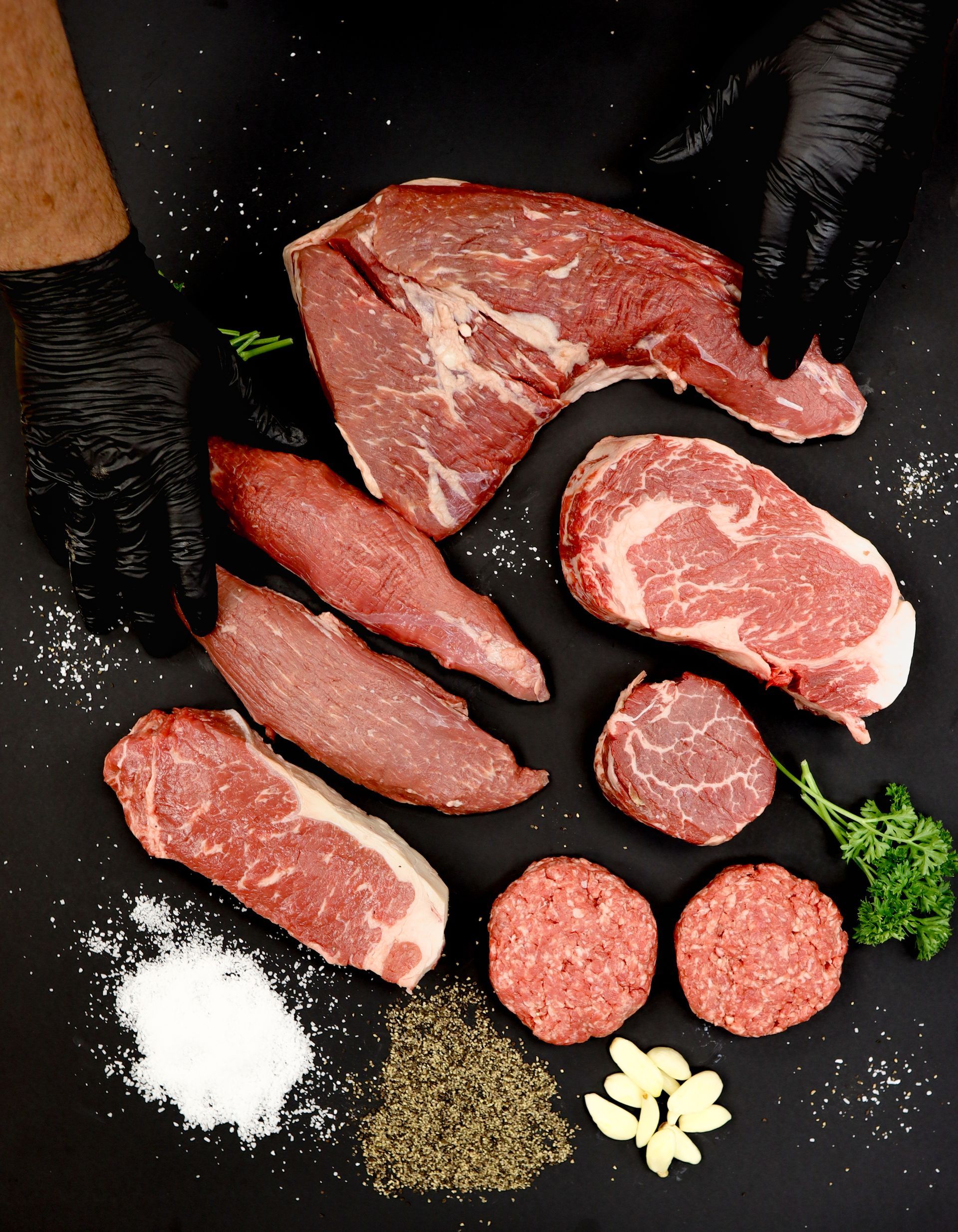 Various raw cuts of meat on a dark surface with salt, pepper, garlic, and parsley, held by gloved hands