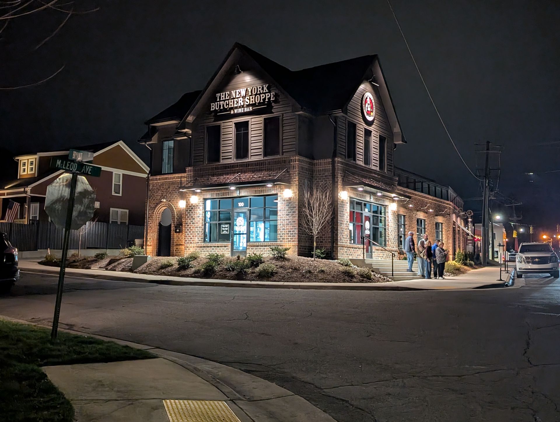 Nighttime view of a lit stone corner building on a quiet street, with storefront windows glowing blue.