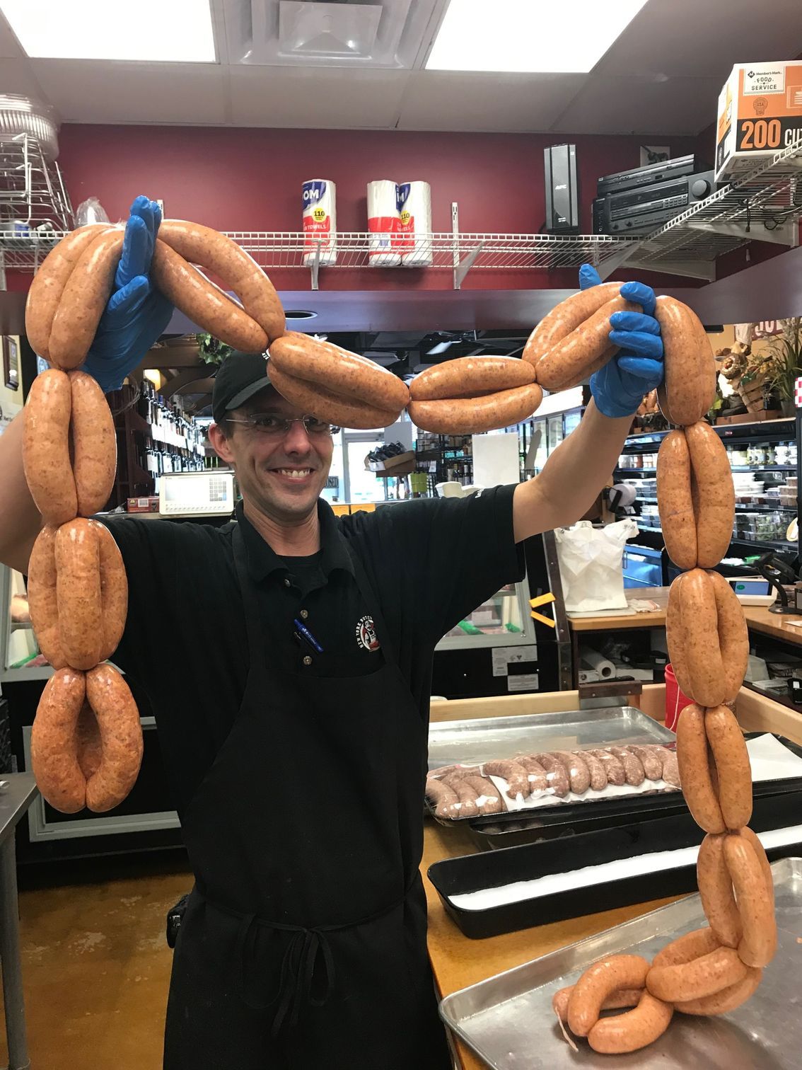 Man in black shirt holds up two linked strands of sausage, smiling. Blue gloves. Shop setting.