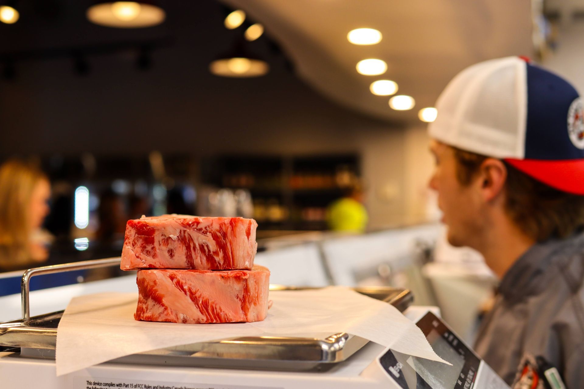 Two raw steaks on a tray in a restaurant, with a person in a cap looking on in the background