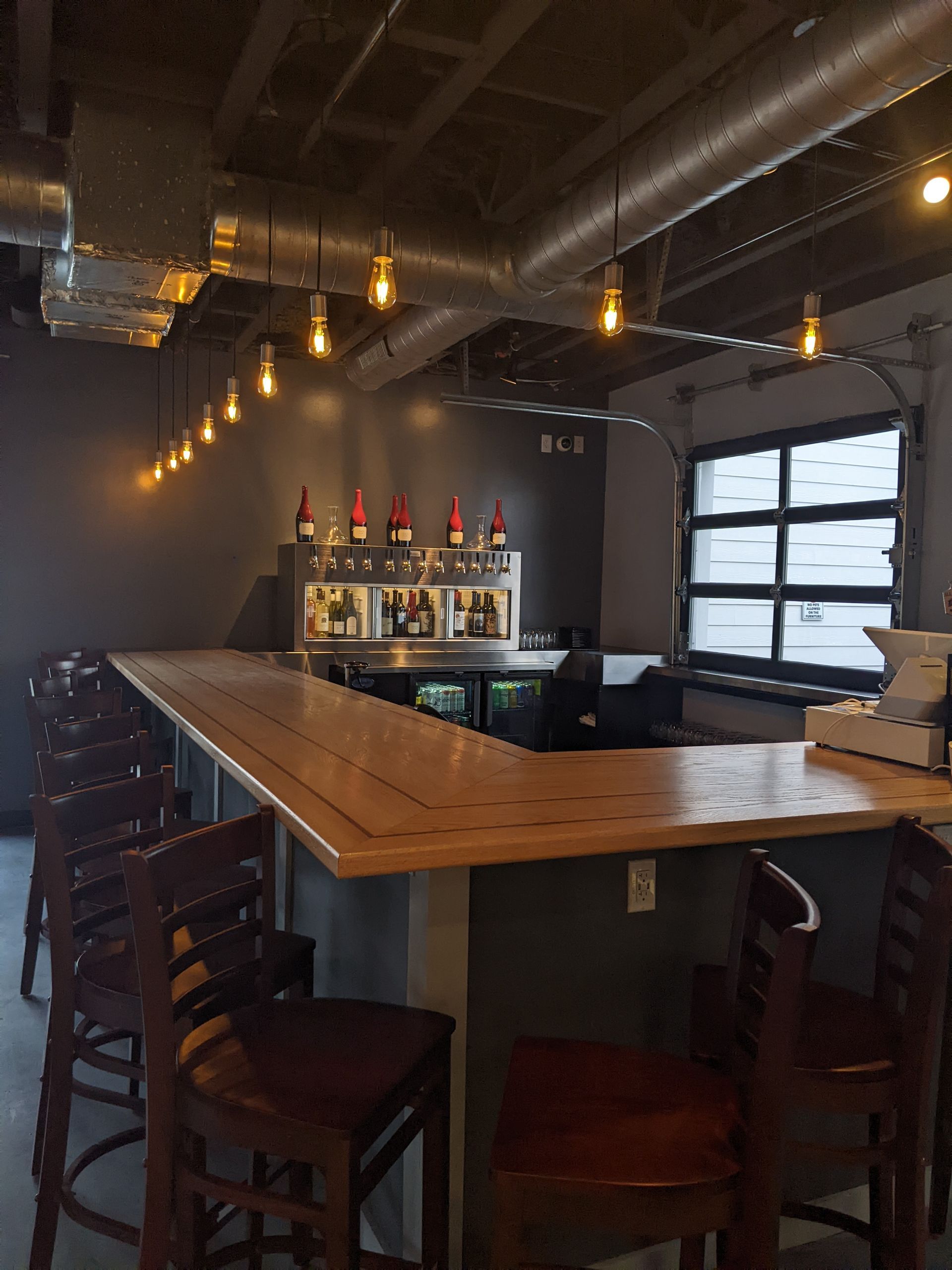Dim industrial-style bar with wooden tables, hanging lights, and a bottle display behind the counter