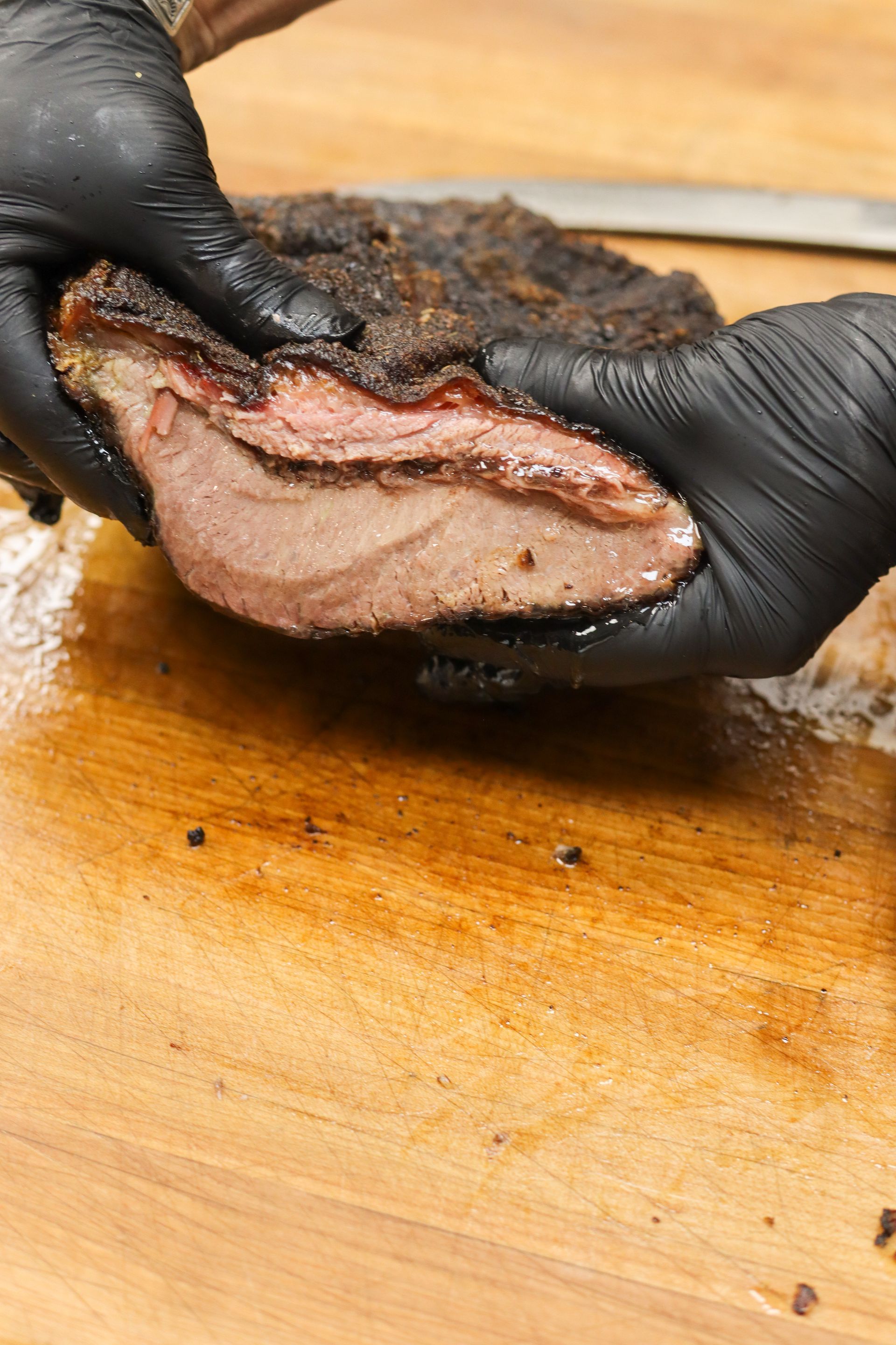 Gloved hands slicing a smoked brisket on a wooden cutting board.