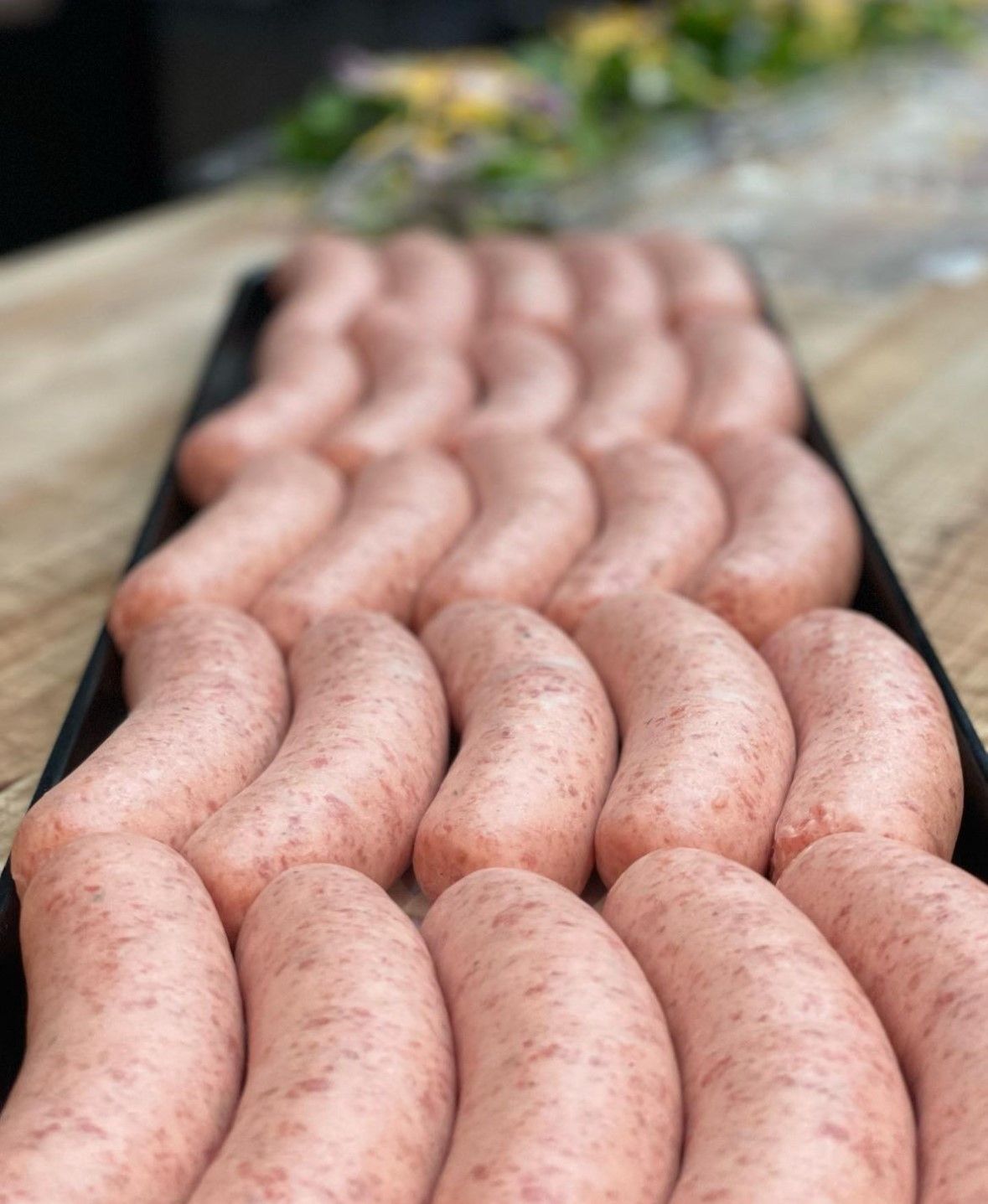 Tray of raw sausages, light pink with visible specks, arranged on a black tray on a wooden surface.