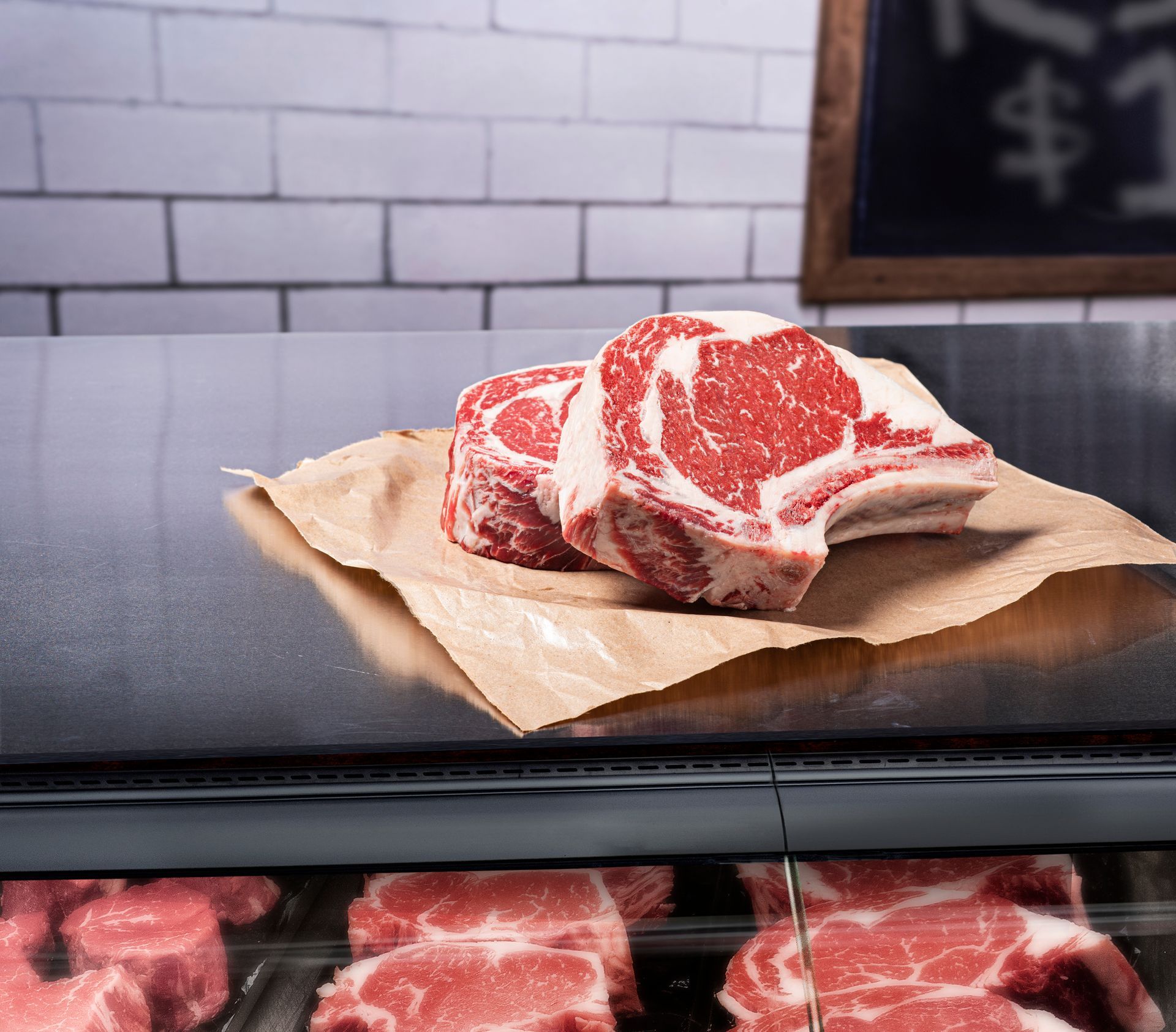 Marbled raw beef steaks on butcher paper at a deli counter, with meat display below.
