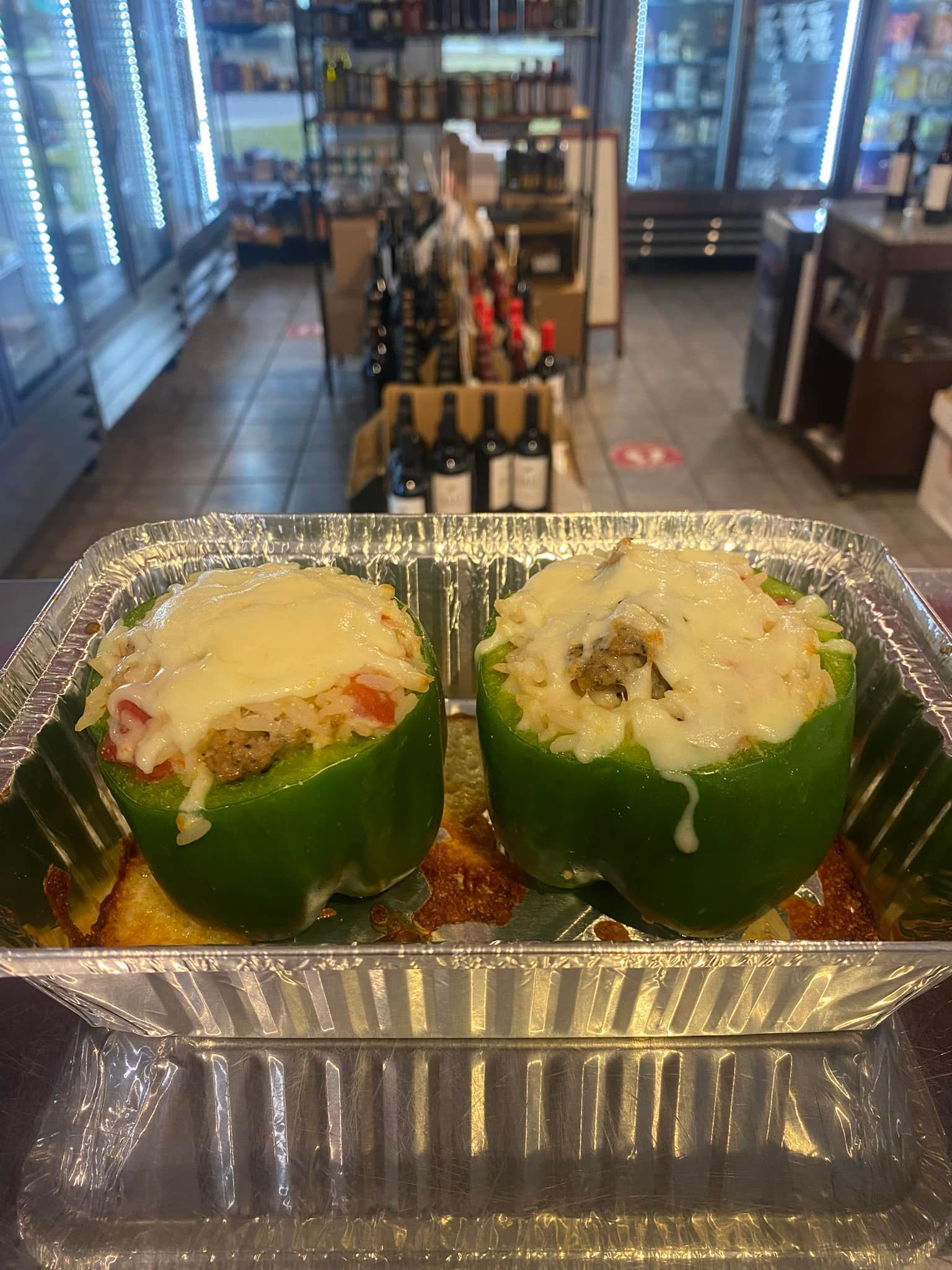 Two stuffed green bell peppers topped with melted cheese in a foil pan, inside a store.