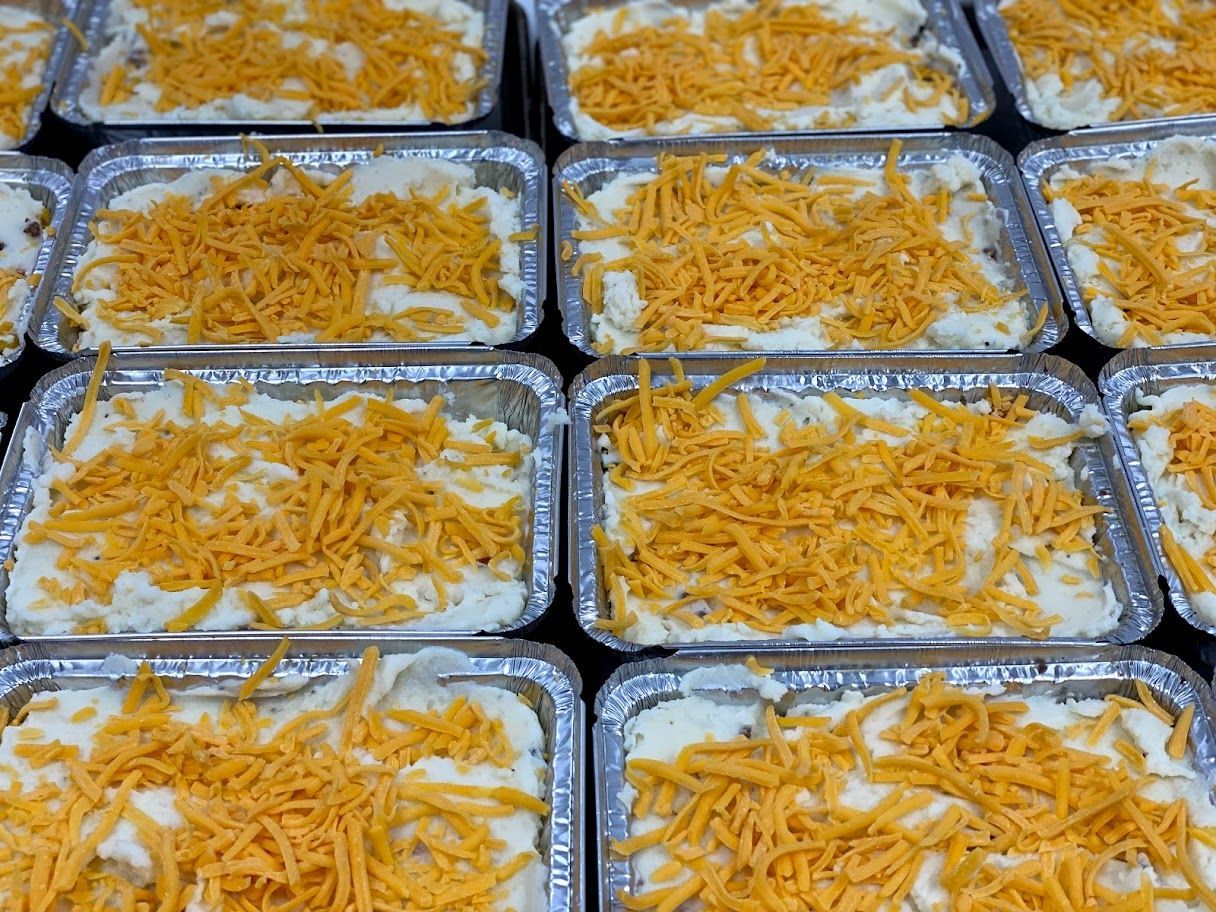 Rows of individual aluminum pans filled with mashed potatoes and topped with shredded cheddar cheese.