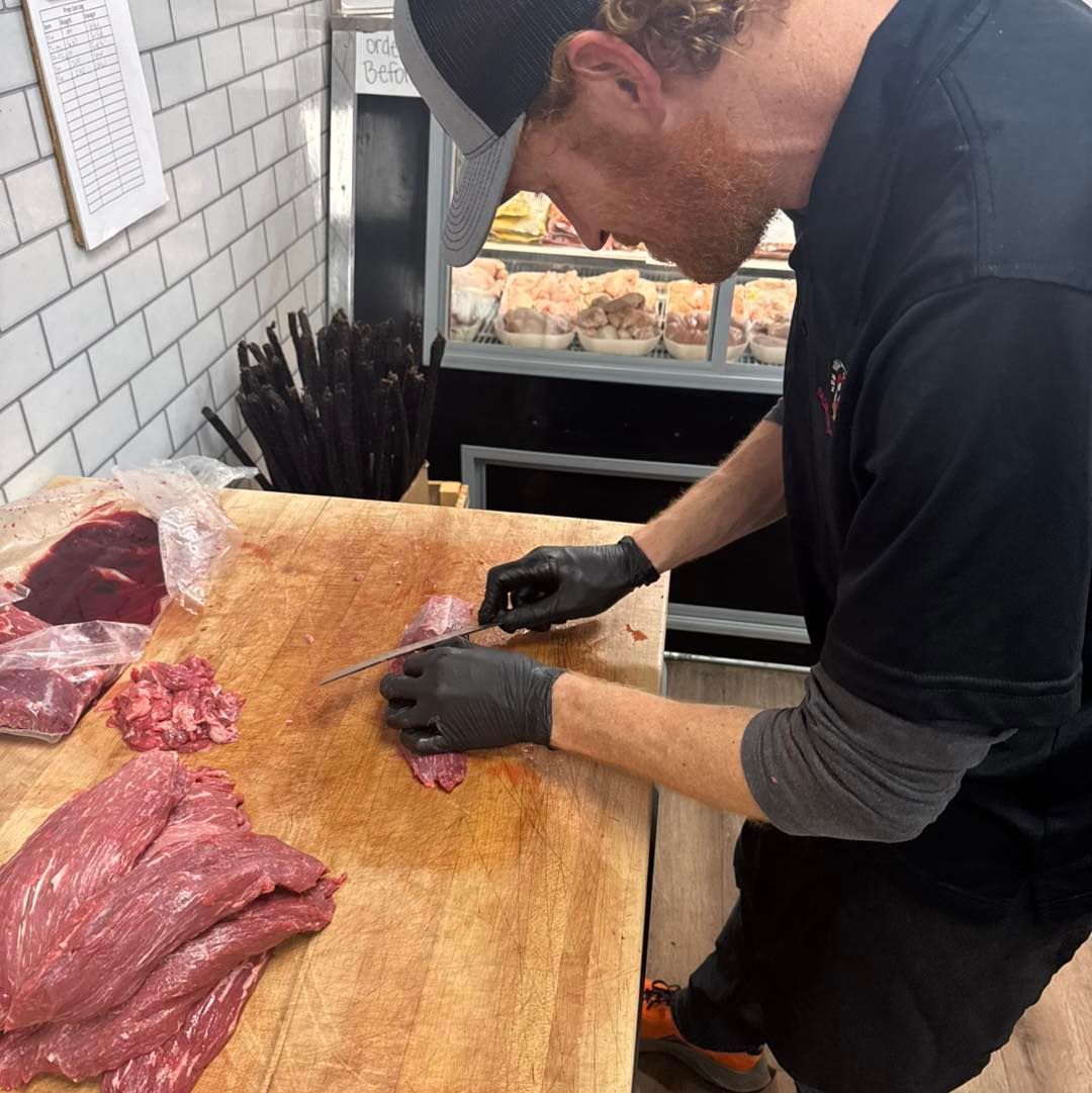 Man in black gloves slicing meat on a wooden cutting board in a butcher shop.