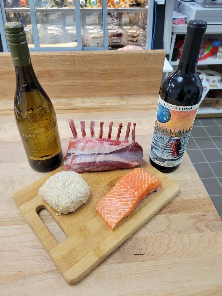 A wooden board with a rack of lamb, salmon, a sesame-crusted ball, and two wine bottles.