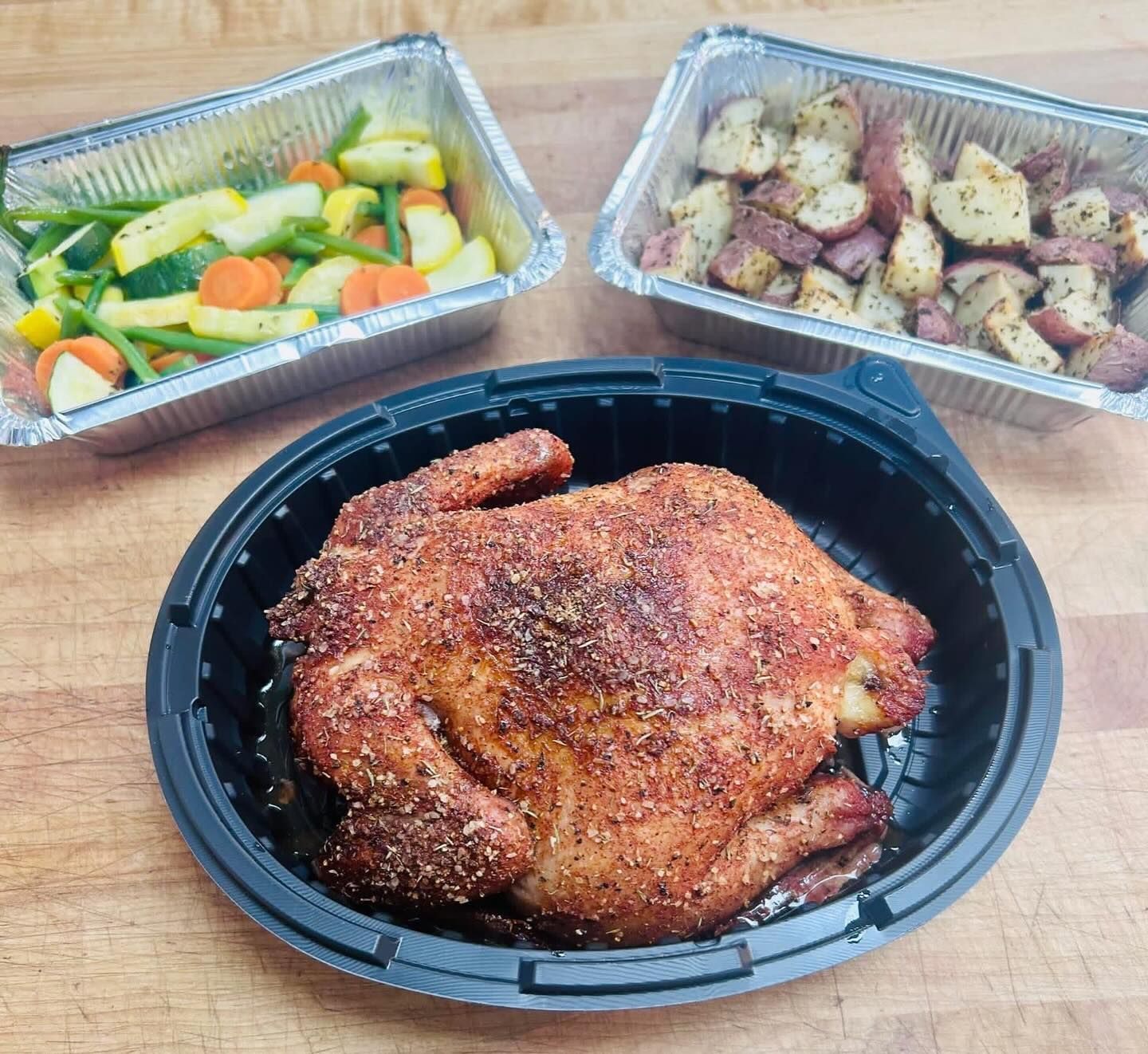 A roasted chicken, with roasted potatoes and mixed vegetables in foil containers, on a wooden table.