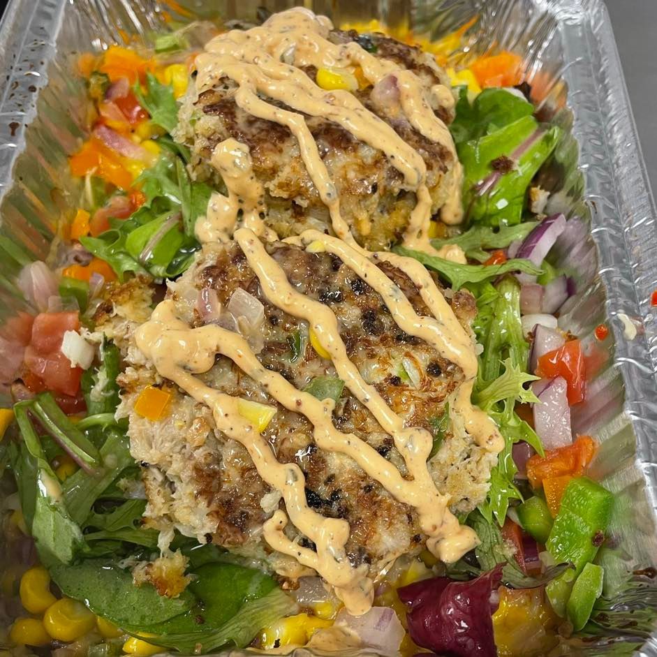 Two crab cakes drizzled with sauce, atop a salad with corn and other colorful vegetables.