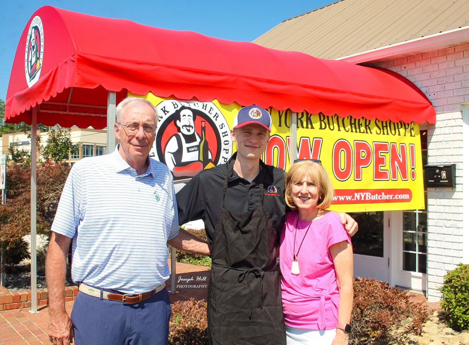 Three people stand in front of a butcher shop with a red awning. The sign says