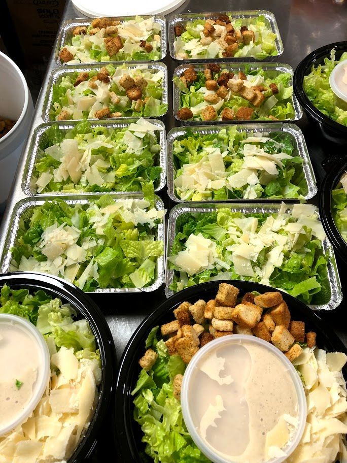 Prepared Caesar salads in black bowls and aluminum trays, with dressing containers.