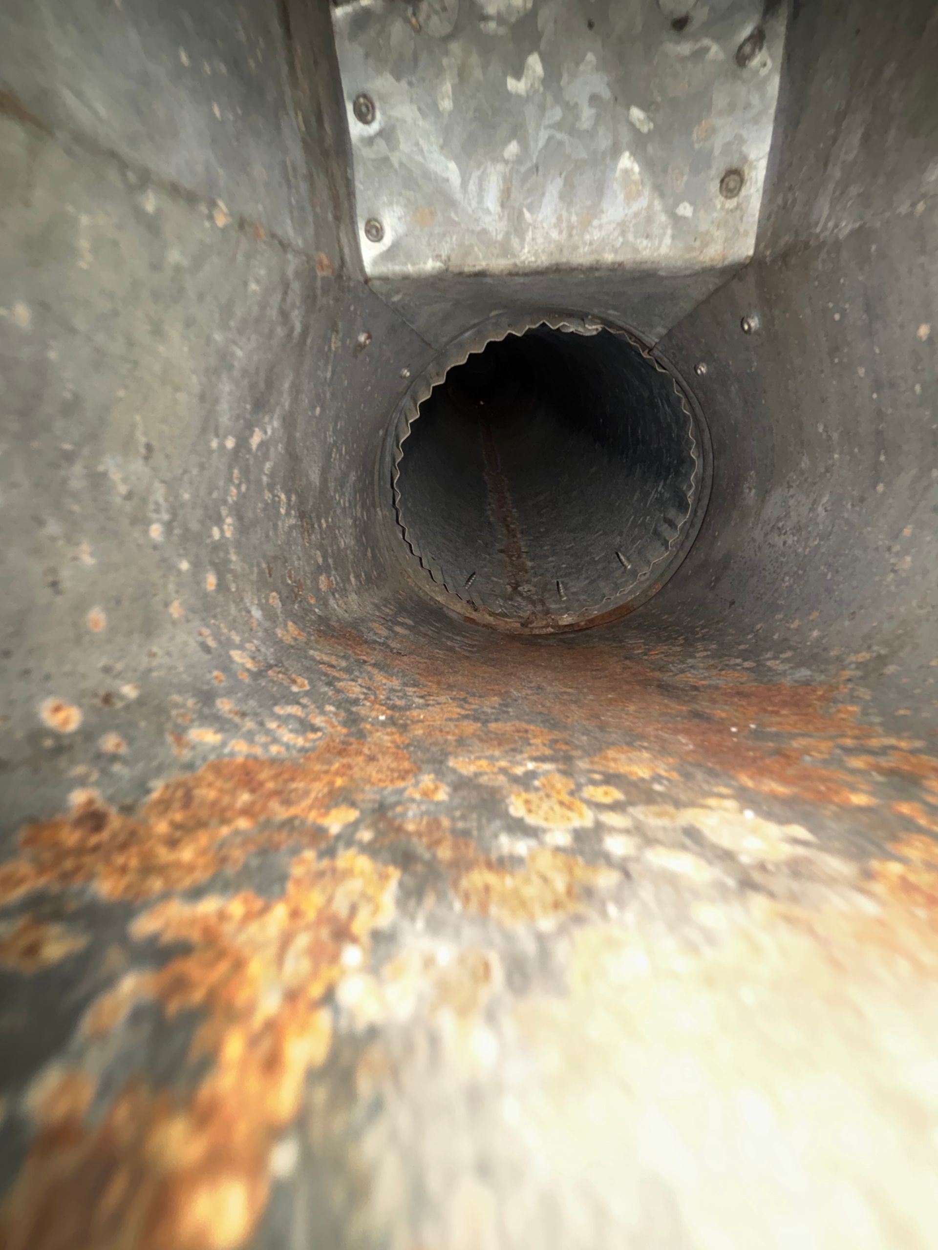 Rusty metal duct interior with a dark circular opening and orange corrosion along the bottom