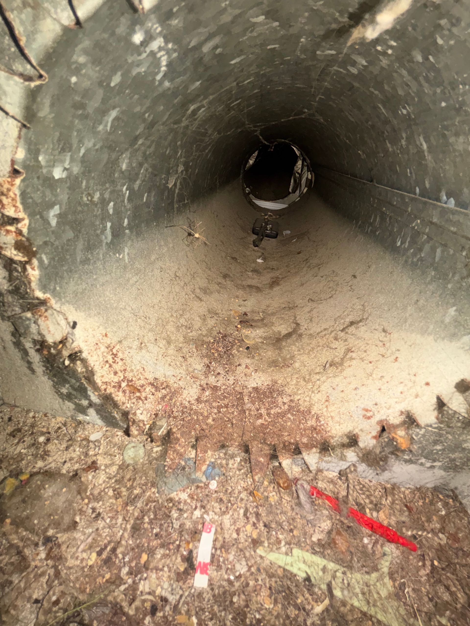 Inside a dusty concrete pipe tunnel with scattered debris and red markings near the opening