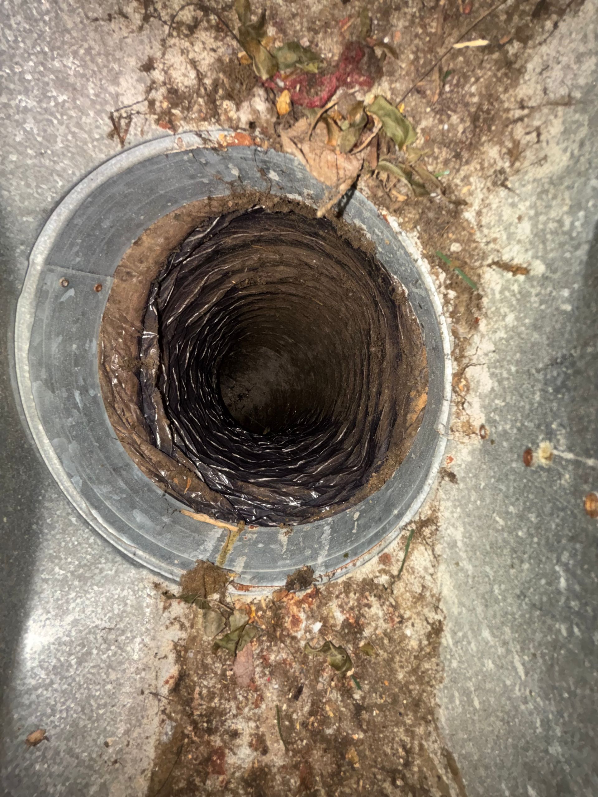 Open metal drain pipe in cracked concrete, with rust and debris around the opening
