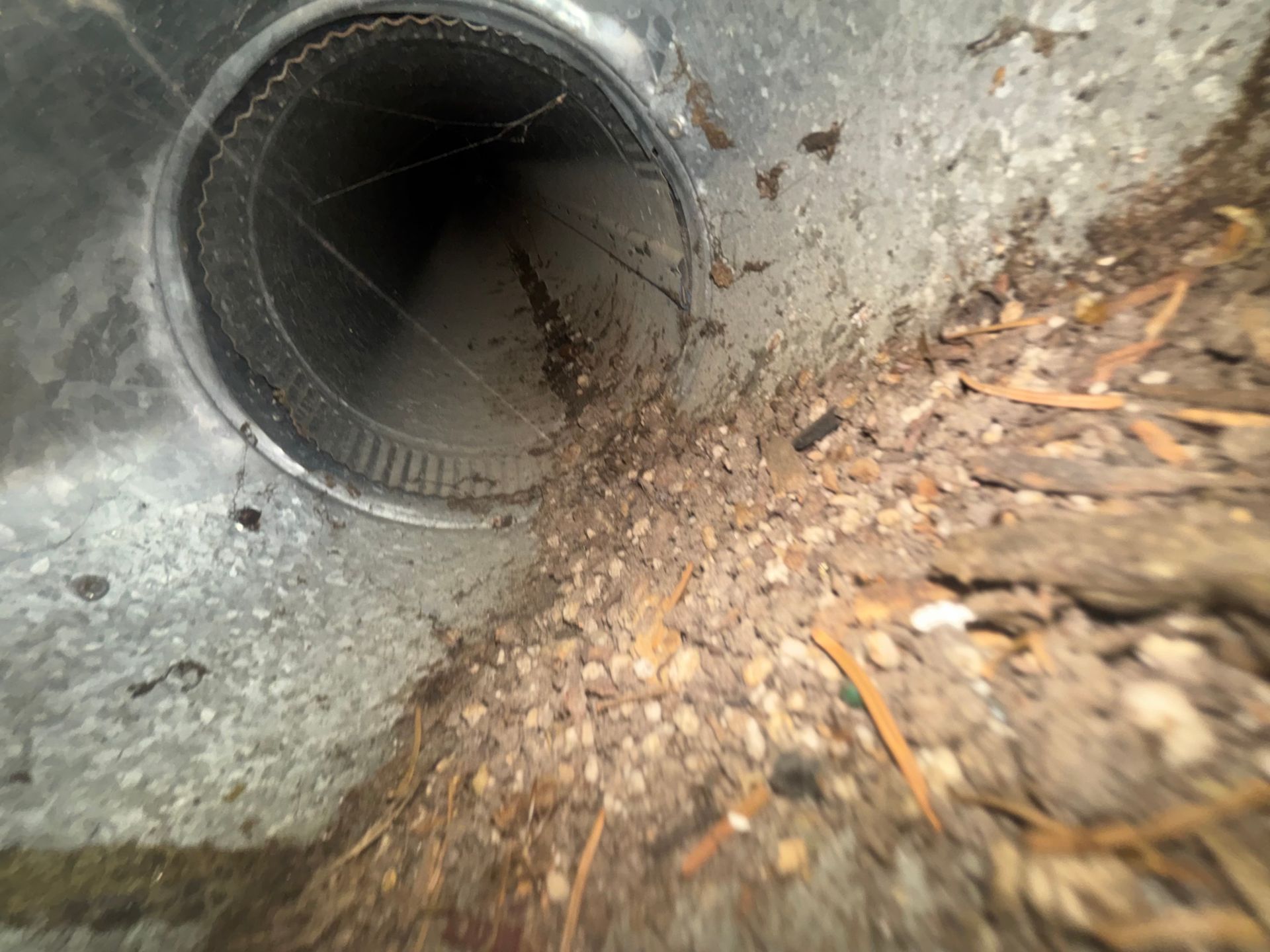 Close-up of a round drain pipe opening in concrete, with dirt and leaves around it.