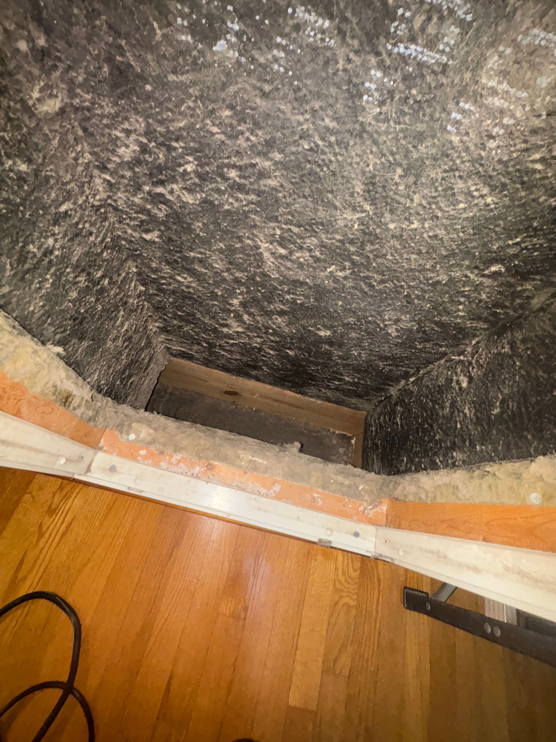 Open floor vent in a hardwood floor beneath a dark insulated ceiling or crawlspace opening