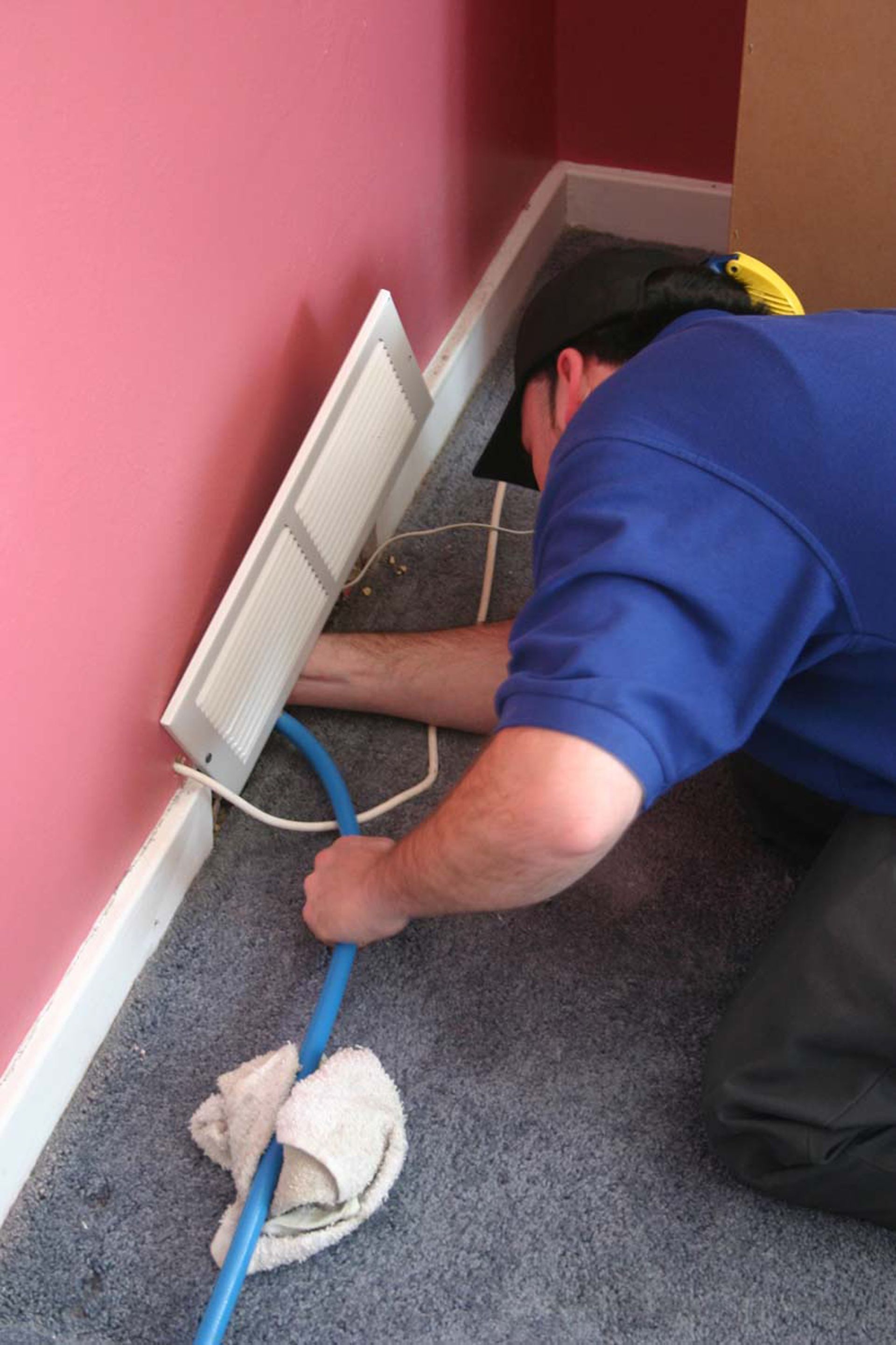 Person kneeling by a wall, cleaning a carpet edge with a blue tool near white vent covers.