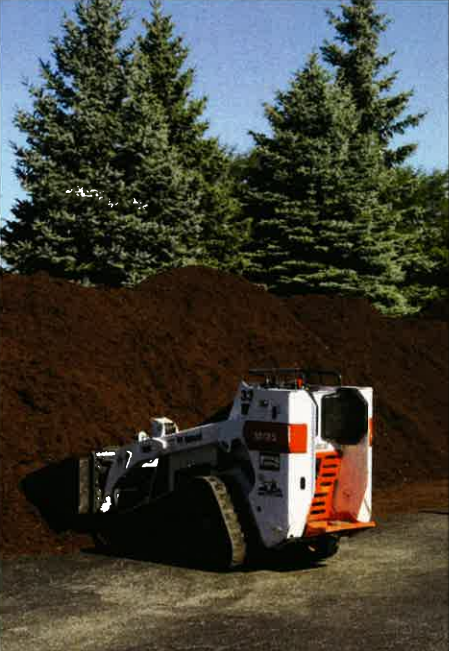 White skid-steer loader with bucket parked in front of a large dirt pile and evergreen trees