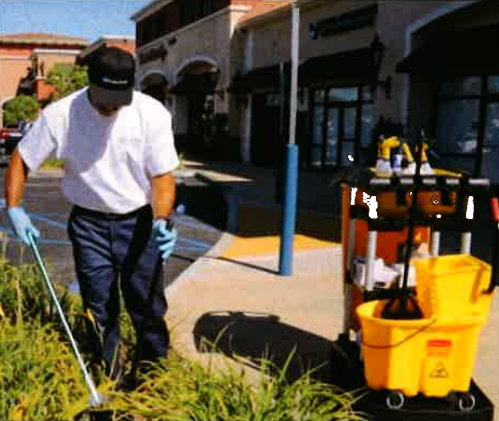 Worker in gloves and mask cleaning landscaping beside a building, with yellow utility cart nearby.