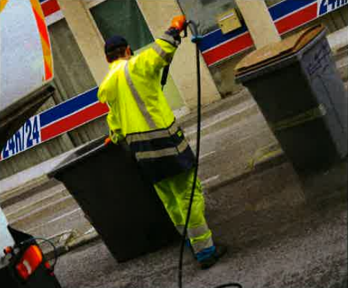 Worker in high-visibility yellow suit handles trash bins beside a parked truck on a wet street.