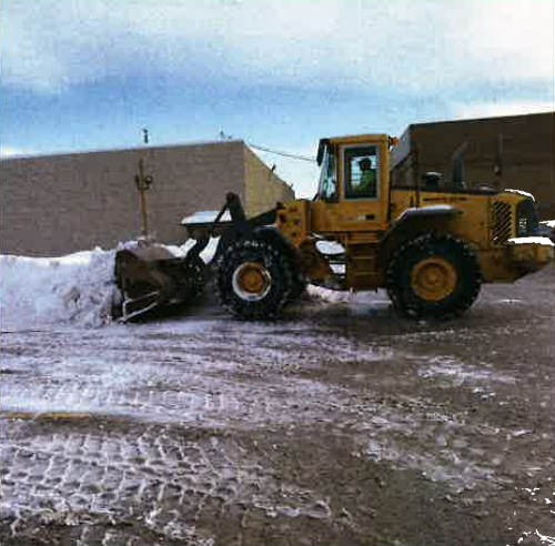 Yellow front loader pushing snow in a snowy lot beside a beige building