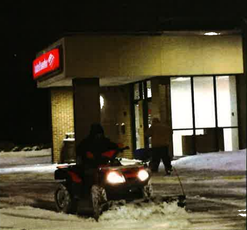 ATV with headlights on in front of a lit storefront at night, on snowy ground.